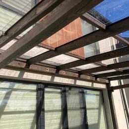 Konstrukcja z belek drewnianych i przezroczystych płyt, prawdopodobnie zadaszenie tarasu lub werandy, widok od dołu, z widocznymi roletami w tle.