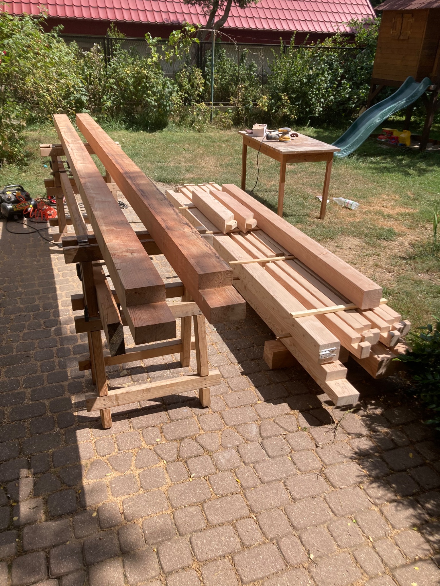 Przygotowane drewniane belki i deski ułożone na kozłach i paletach na brukowanym podwórku, narzędzia i stół w tle, widoczna zieleń ogrodu i domek ze zjeżdżalnią.