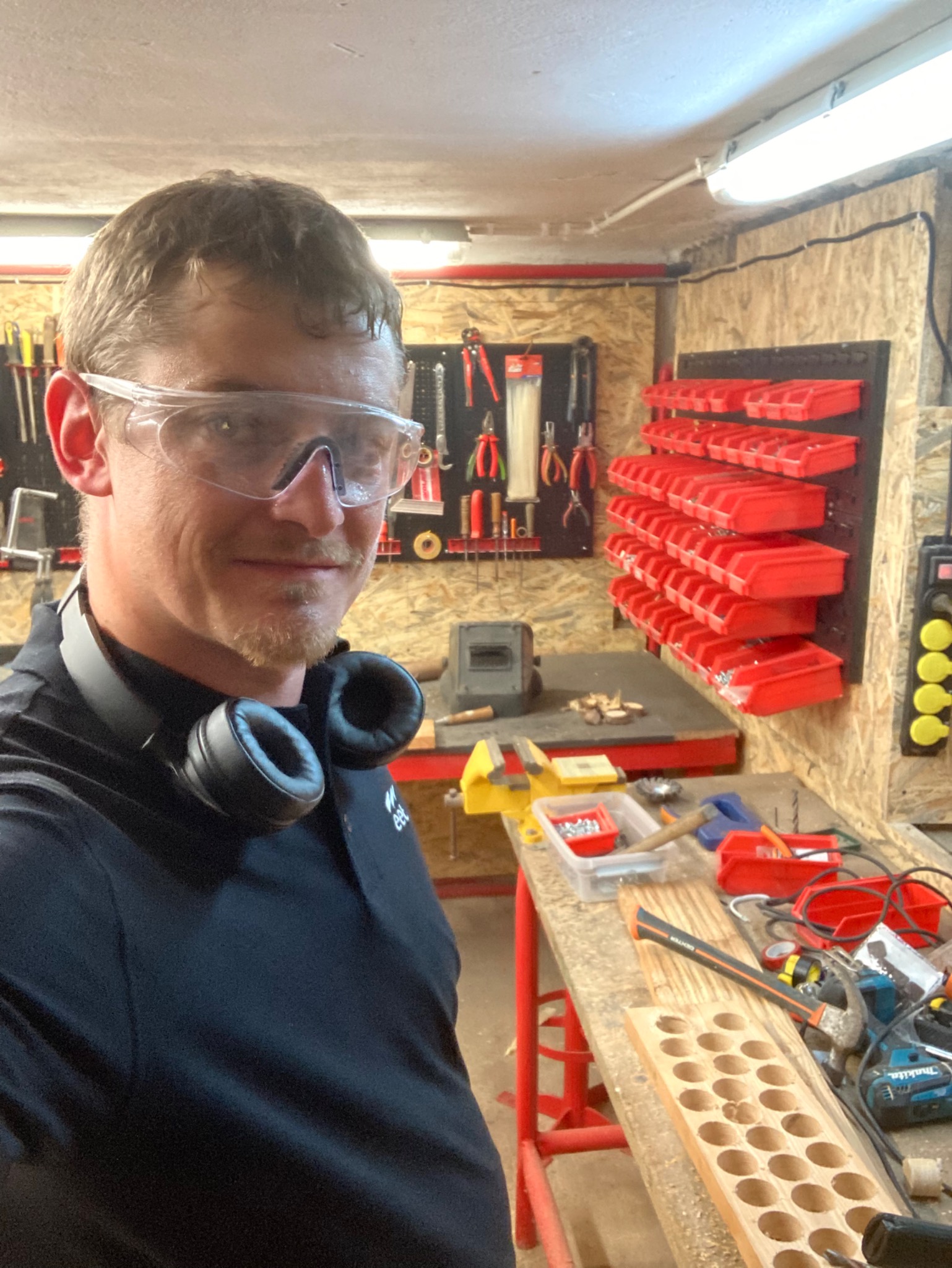 Autoportret rzemieślnika w okularach ochronnych, z słuchawkami na szyi, w warsztacie z narzędziami i deską z wywierconymi otworami.