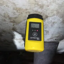 Żółty wilgotnościomierz Trotec BM18, z włączonym wyświetlaczem, przyłożony do szarej, nierównej powierzchni, prawdopodobnie ściany lub stropu w piwnicy.