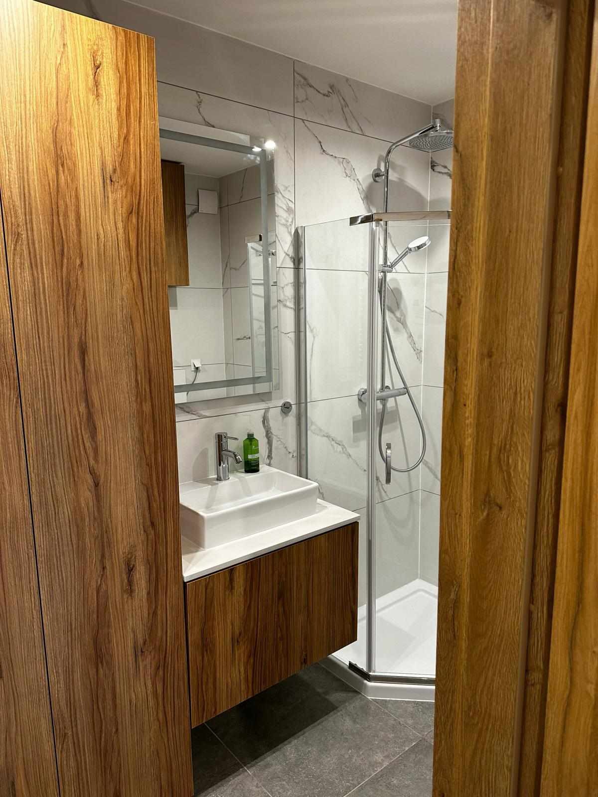 Nowoczesna łazienka z kabiną prysznicową, umywalką nablatową na szafce z drewnianym fornirem i lustrem w metalowej ramie, wykończona płytkami imitującymi marmur.