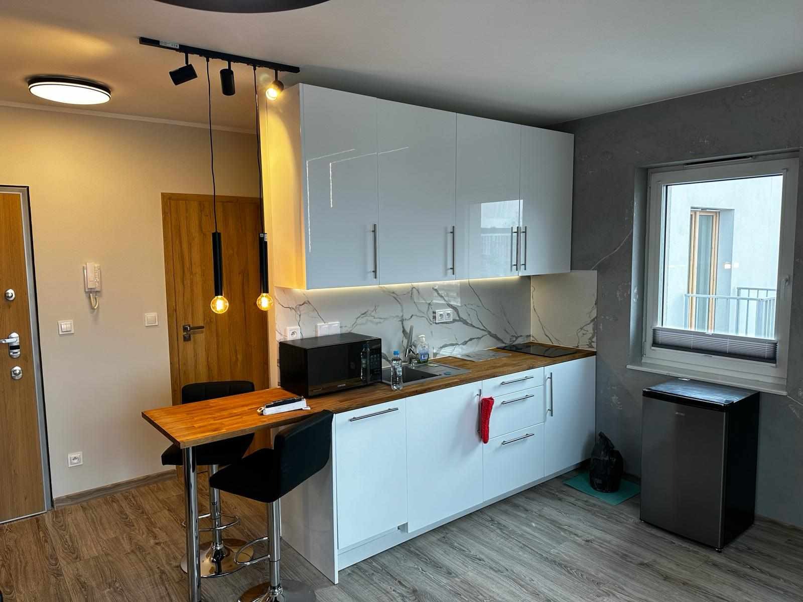 Nowoczesna kuchnia z białymi, lakierowanymi szafkami, blatem imitującym drewno, marmurowym backsplashiem i barkiem z drewnianym blatem oraz czarnymi hokerami, oświetlona wiszącymi lampami...