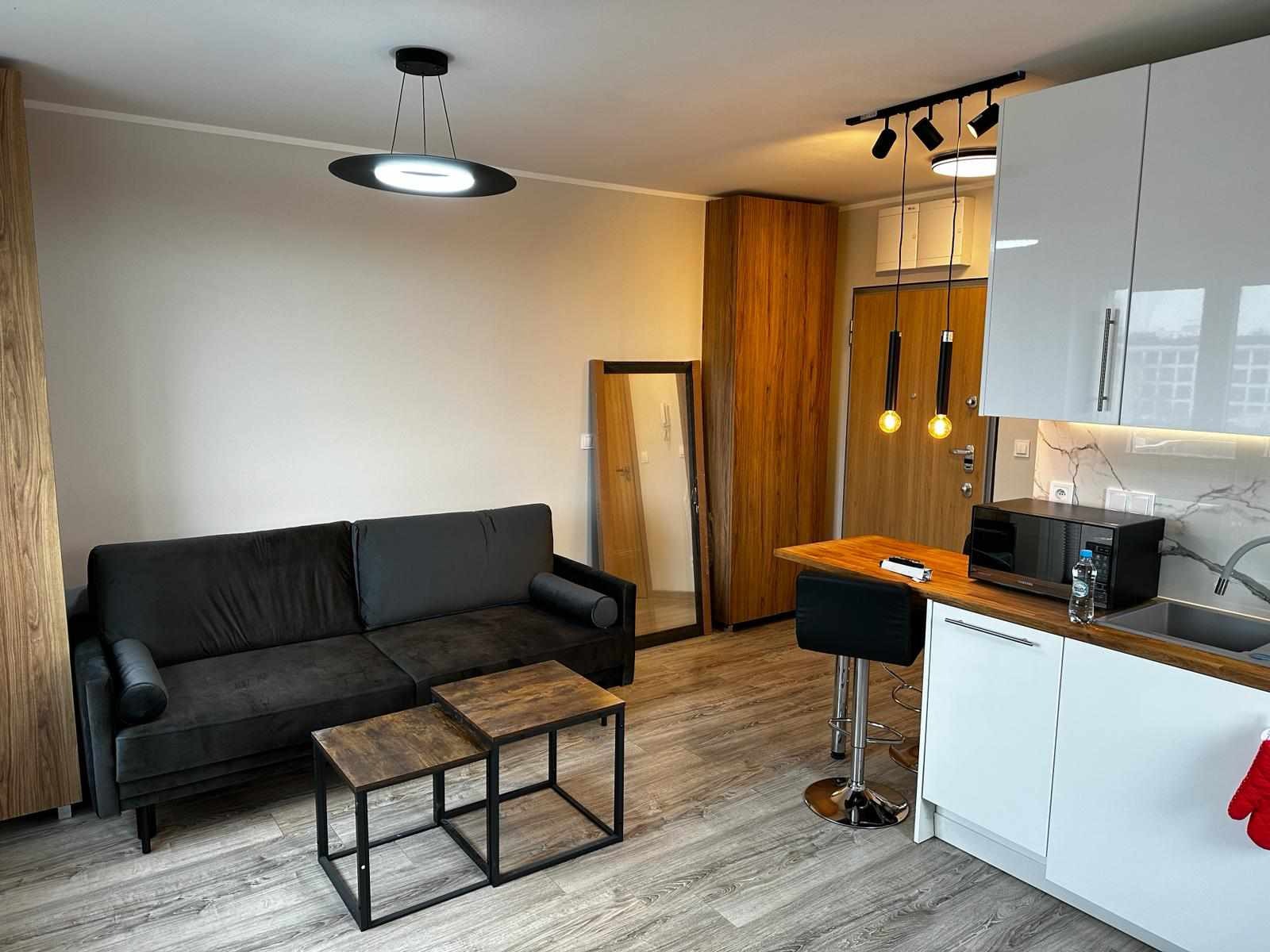 Wnętrze kawalerki z aneksem kuchennym: ciemnoszara sofa, drewniane stoliki kawowe, wysoki taboret barowy, białe szafki kuchenne z marmurowym backsplash, drewniana szafa, lustro w czarnej ramie...