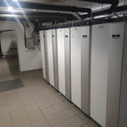 Asterm - Instalacje Sanitarne - Szereg białych i szarych jednostek PC10, PC9, PC8, PC7, PC6, PC5, PC4, PC3, PC2, PC1, prawdopodobnie pomp ciepła, zainstalowanych w piwnicy lub pomieszczeniu technicznym z widocznymi rurami...