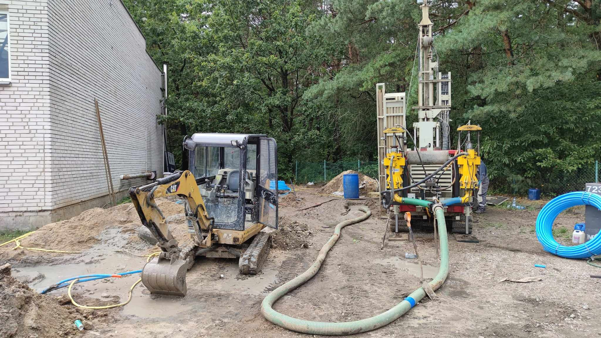 Żółty minikoparka CAT i wiertnica na błotnistym placu budowy, z widocznymi niebieskimi rurami i fragmentem budynku w tle.