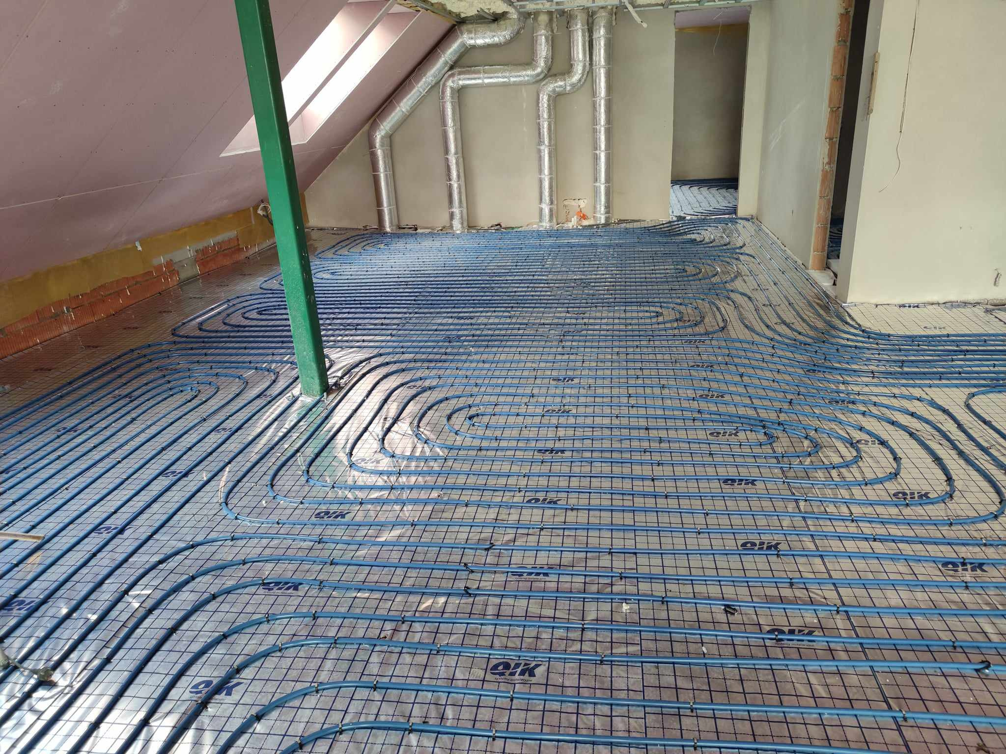 Niebieskie rury ogrzewania podłogowego ułożone na siatce w surowym wnętrzu poddasza, widoczne przewody wentylacyjne pod sufitem i okno dachowe.