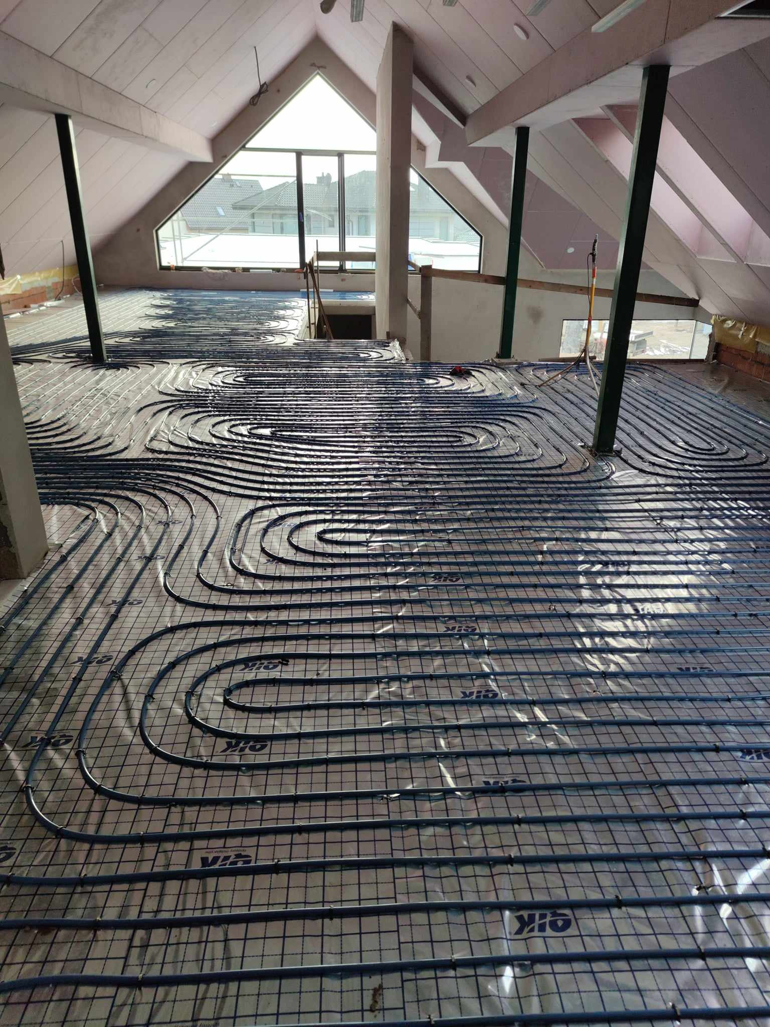 Montaż ogrzewania podłogowego w poddaszu: rury grzewcze ułożone na folii z siatką, widoczne okno dachowe i konstrukcja dachu.