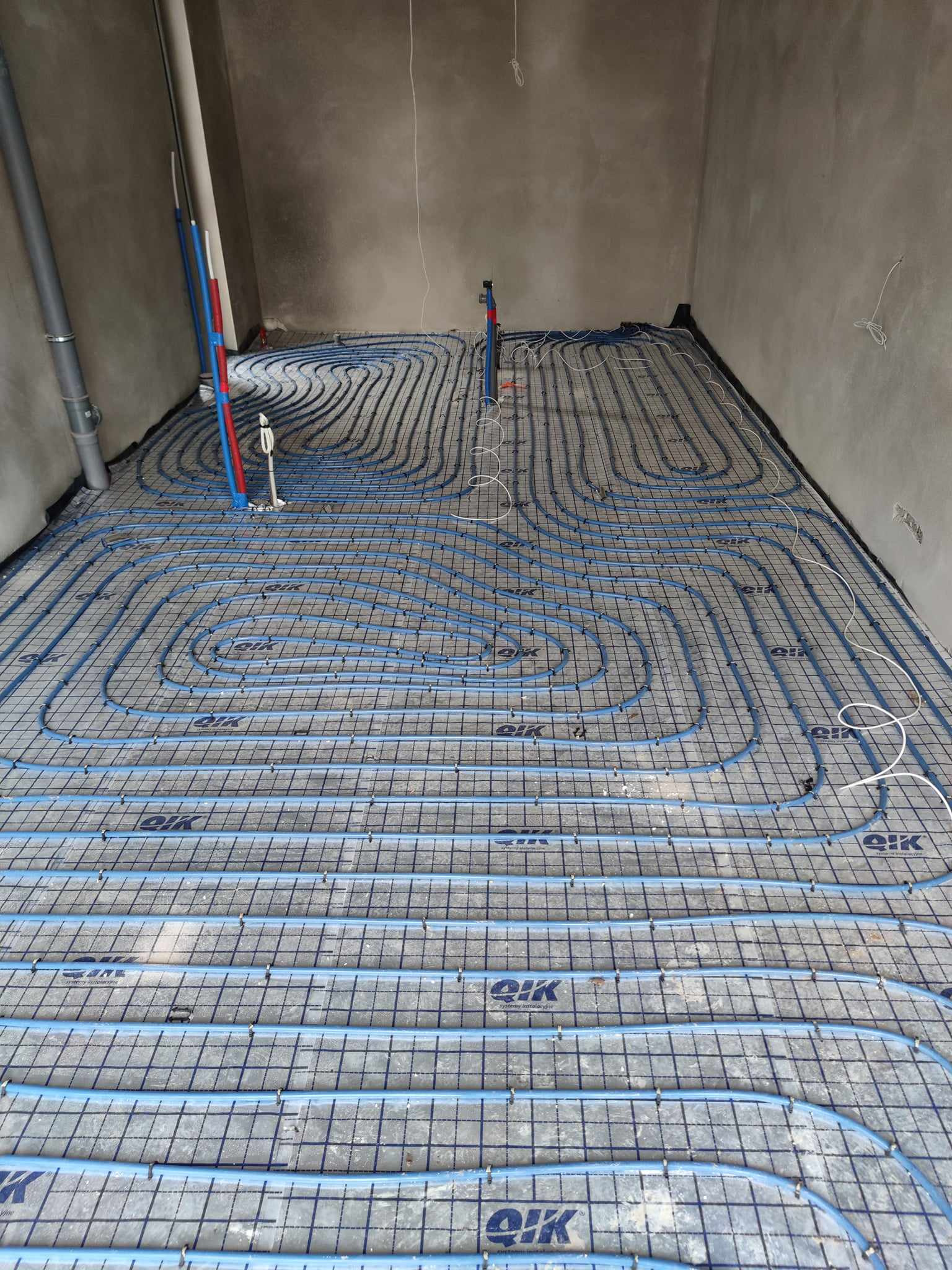 Rozłożone rury ogrzewania podłogowego w surowym pomieszczeniu, widoczne przyłącza pionowe rur w kolorze czerwonym i niebieskim, mata z nadrukowanym logo producenta.