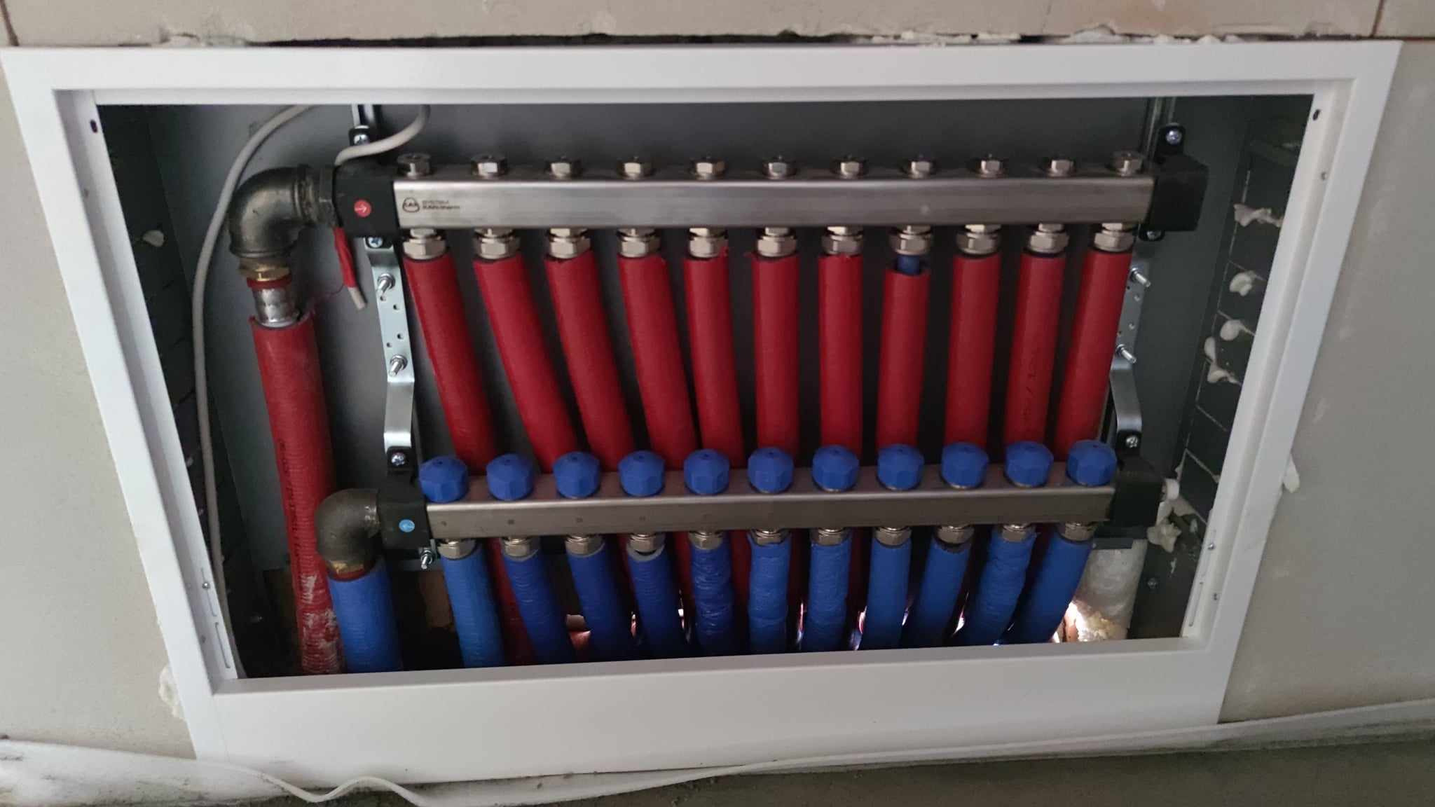 Rozdzielacz ogrzewania podłogowego z czerwonymi i niebieskimi rurami w białej obudowie, widoczne przyłącza i zawory regulacyjne.