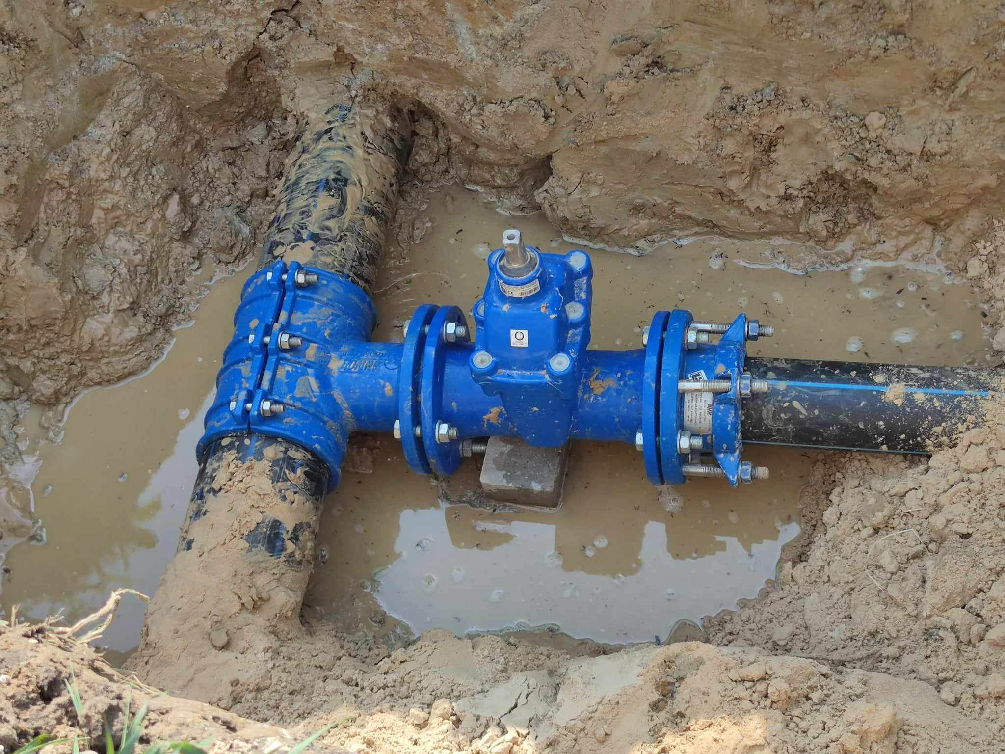 Niebieski zawór z kołnierzami na czarnej rurze w wykopie wypełnionym błotnistą wodą; widoczny fragment rury odchodzącej w lewo, całość w ziemnym wykopie.
