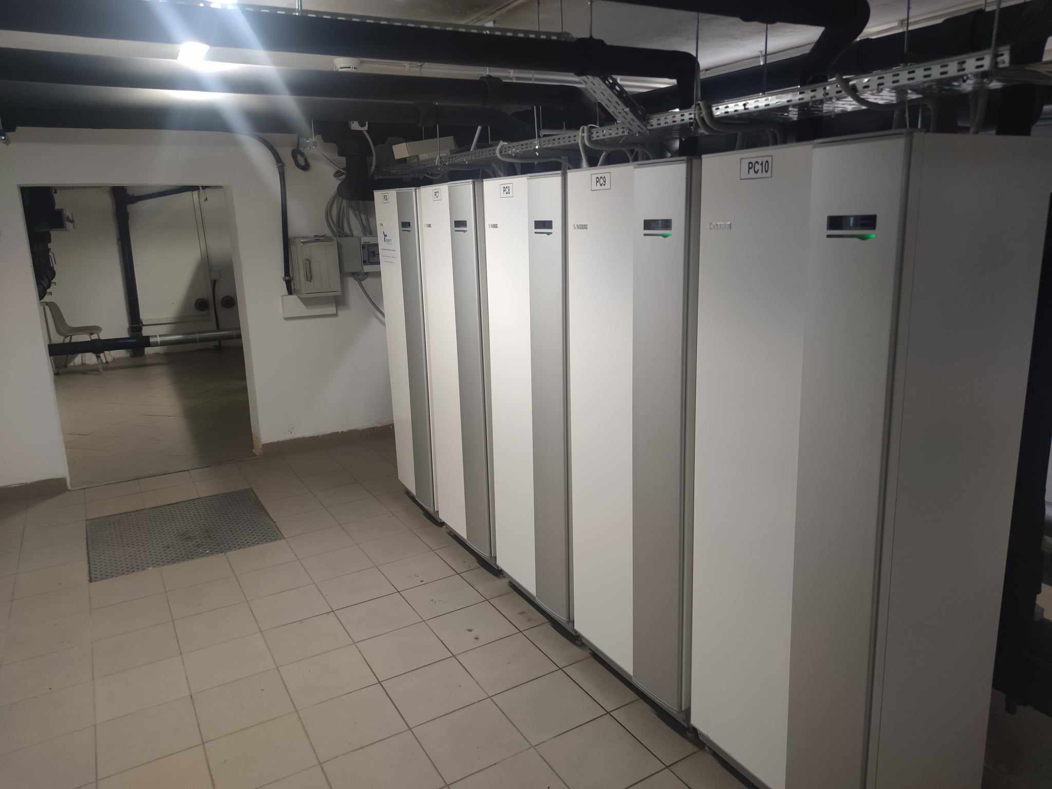 Szereg białych i szarych jednostek PC10, PC9, PC8, PC7, PC6, PC5, PC4, PC3, PC2, PC1, prawdopodobnie pomp ciepła, zainstalowanych w piwnicy lub pomieszczeniu technicznym z widocznymi rurami...