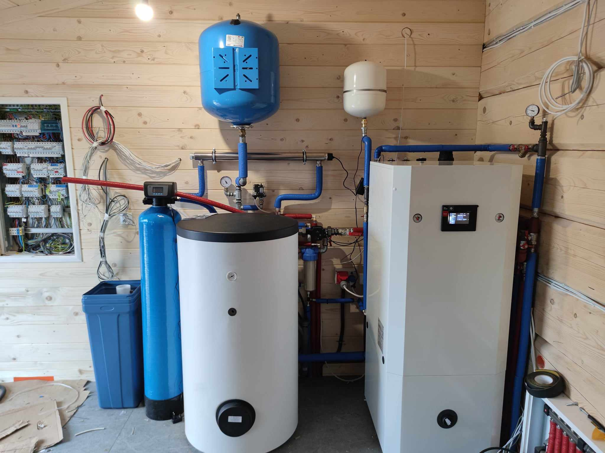Kompletna instalacja grzewcza w kotłowni z drewnianą boazerią, obejmująca biały zasobnik ciepłej wody, niebieski hydrofor, filtr wody i rozdzielnicę elektryczną z licznymi przewodami.