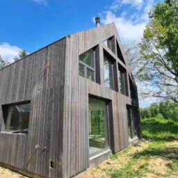 Nowoczesny dom z elewacją z ciemnego drewna, duże okna, widok z zewnątrz w słoneczny dzień, trawa i drzewa w tle.