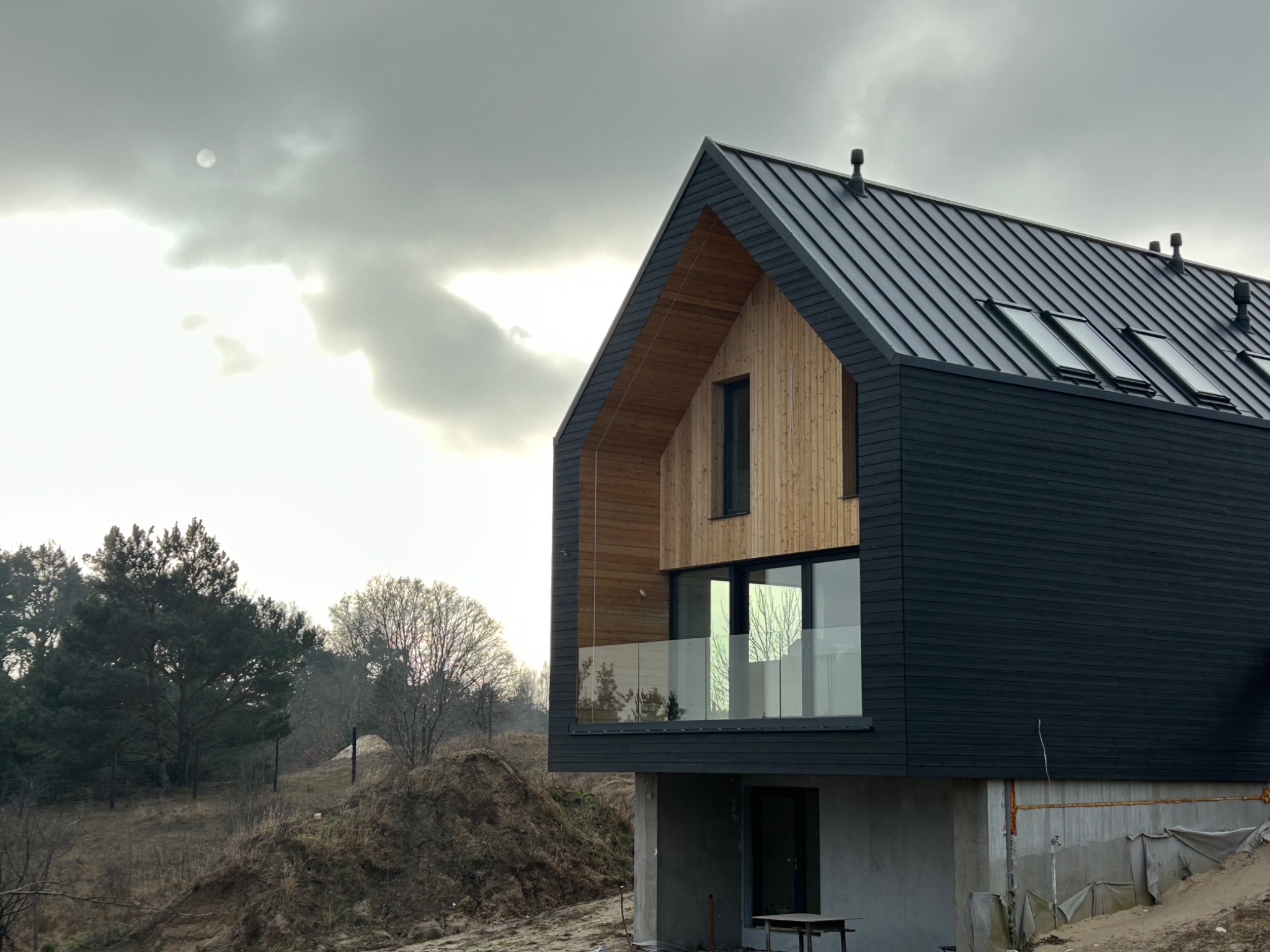 Nowoczesny dom z ciemną elewacją z drewnianymi akcentami, szklaną balustradą balkonu i metalowym dachem w pochmurny dzień.