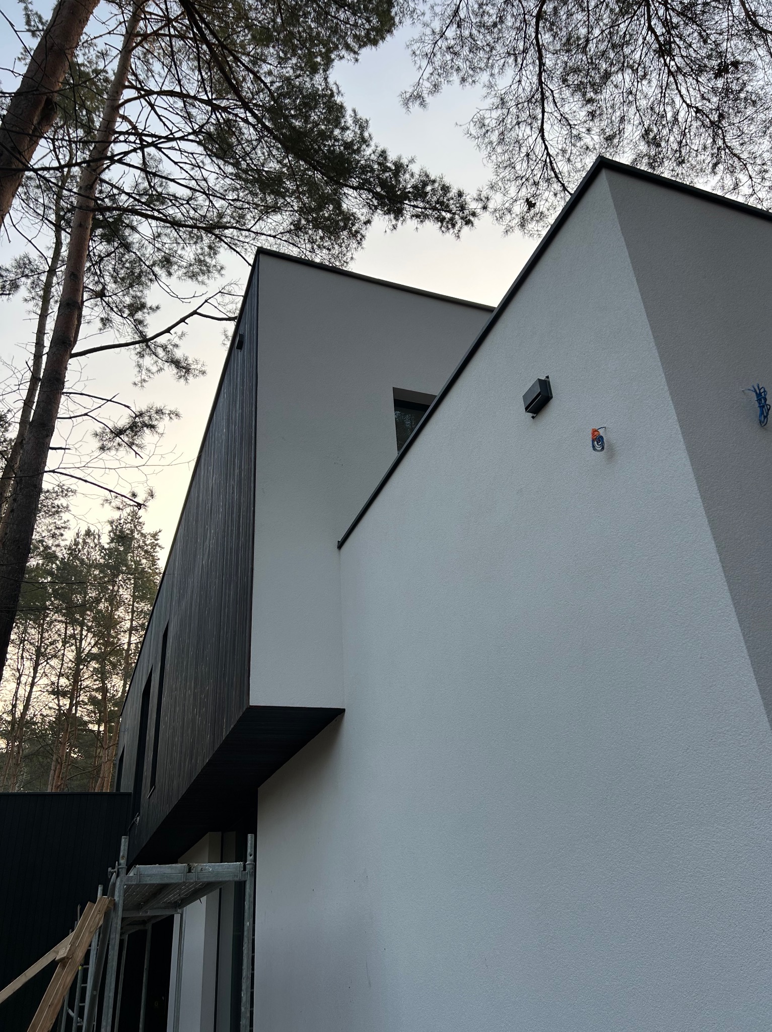 Nowoczesny dom z białą elewacją i elementami z ciemnego drewna, widoczne rusztowanie i fragmenty instalacji elektrycznej na ścianie.