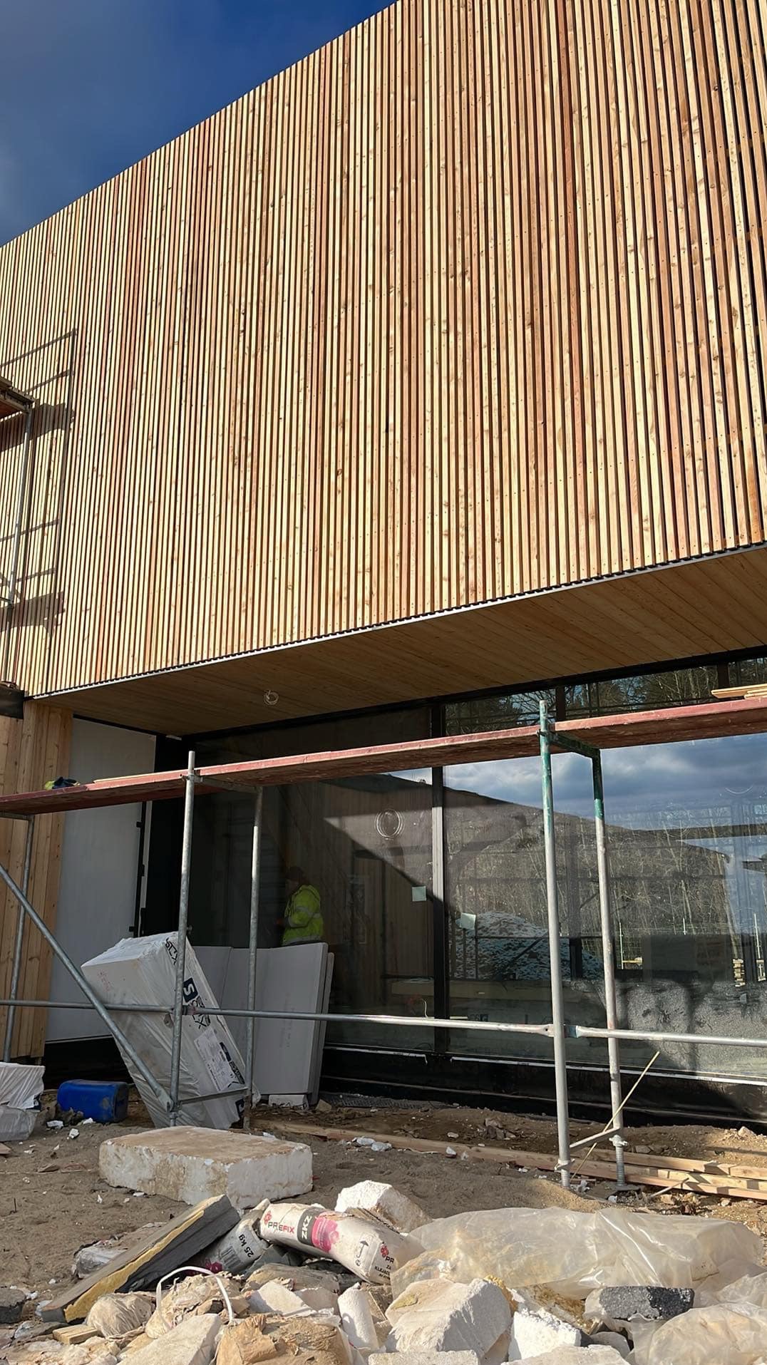 Nowoczesny budynek z elewacją z pionowych desek, fragment rusztowania i materiały budowlane na pierwszym planie, widoczny pracownik w odblaskowej kamizelce wewnątrz budynku.