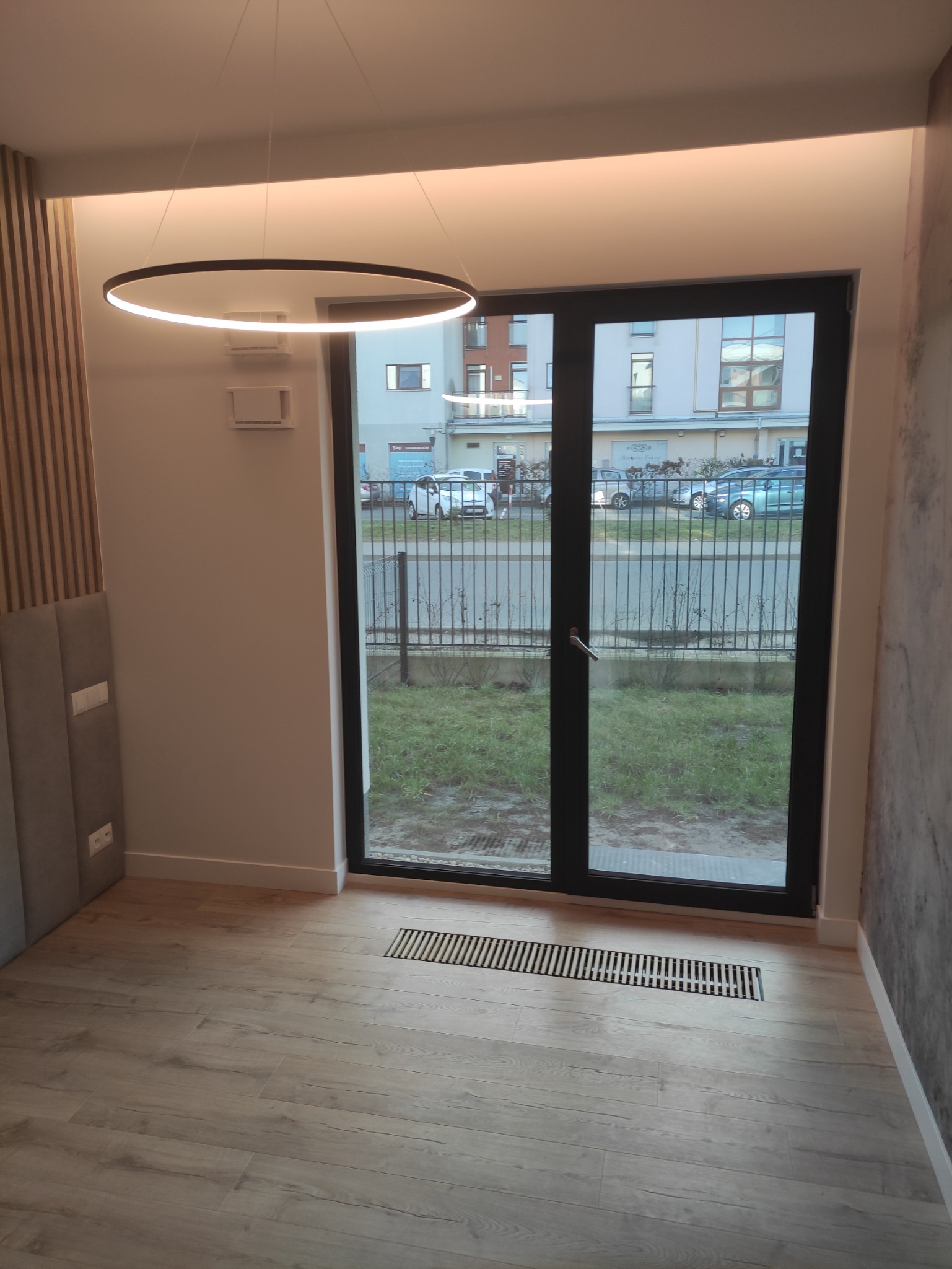 Jasne wnętrze z drewnopodobną podłogą, okrągłą lampą sufitową LED i widokiem na zewnątrz przez czarne drzwi balkonowe.