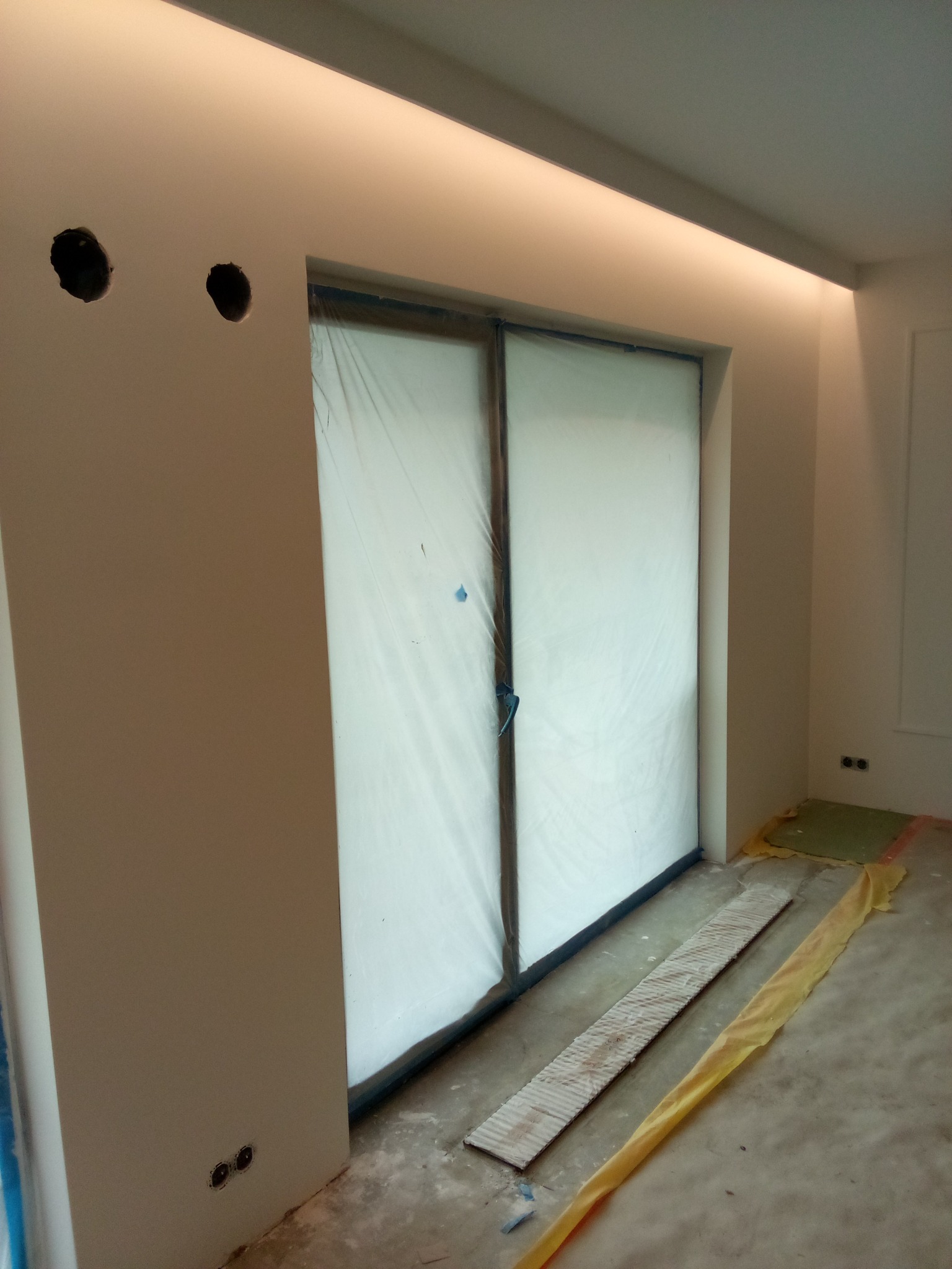 Przygotowanie do prac wykończeniowych: zabezpieczone folią drzwi przesuwne, widoczne otwory na gniazdka elektryczne i listwa LED pod sufitem.