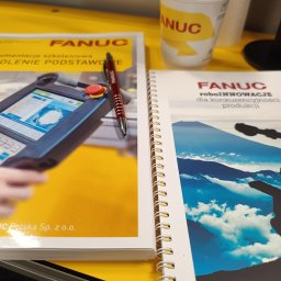 STUDIO AUTOMATYKI PRZEMYSŁOWEJ KOMERCYJNEJ I DOMOWEJ SŁAWOMIR KORCZYŃSKI - Dokumentacja szkoleniowa i notatnik z robotem przemysłowym Fanuc oraz długopis i kubek z logo Fanuc na żółtym tle.