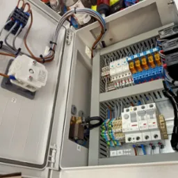 Otwarta szafa sterownicza z widocznymi elementami elektrycznymi: styczniki, bezpieczniki, przewody w peszlach, złączki, opisane etykietami, z narzędziami i akcesoriami na górze.