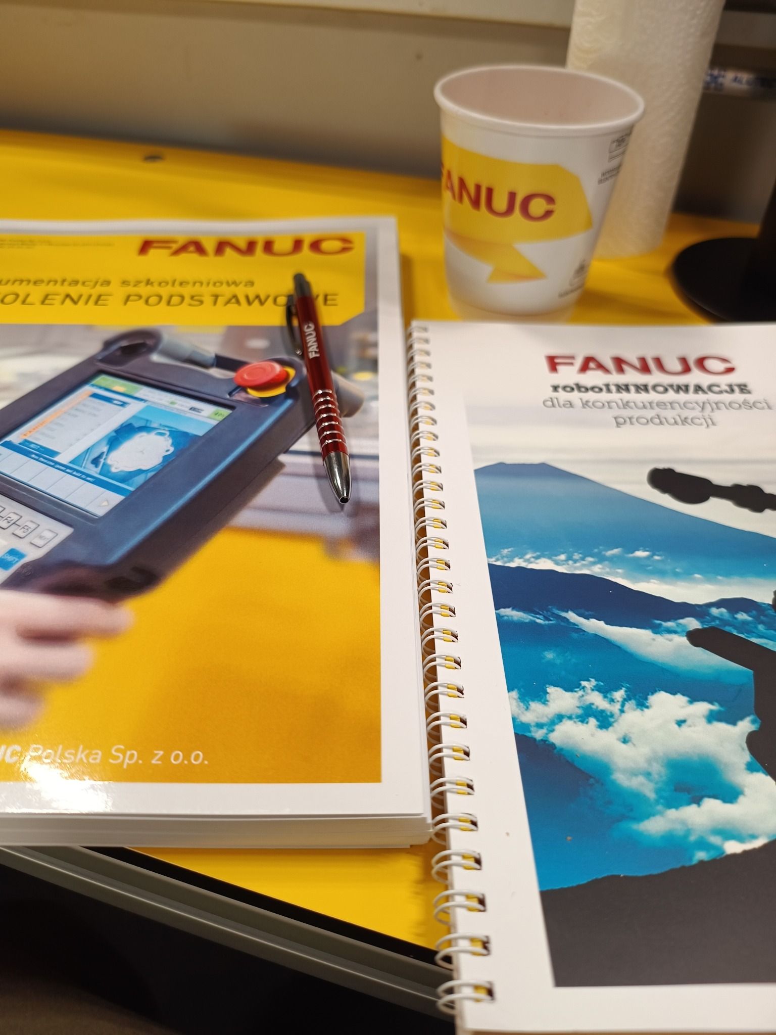 Dokumentacja szkoleniowa i notatnik z robotem przemysłowym Fanuc oraz długopis i kubek z logo Fanuc na żółtym tle.