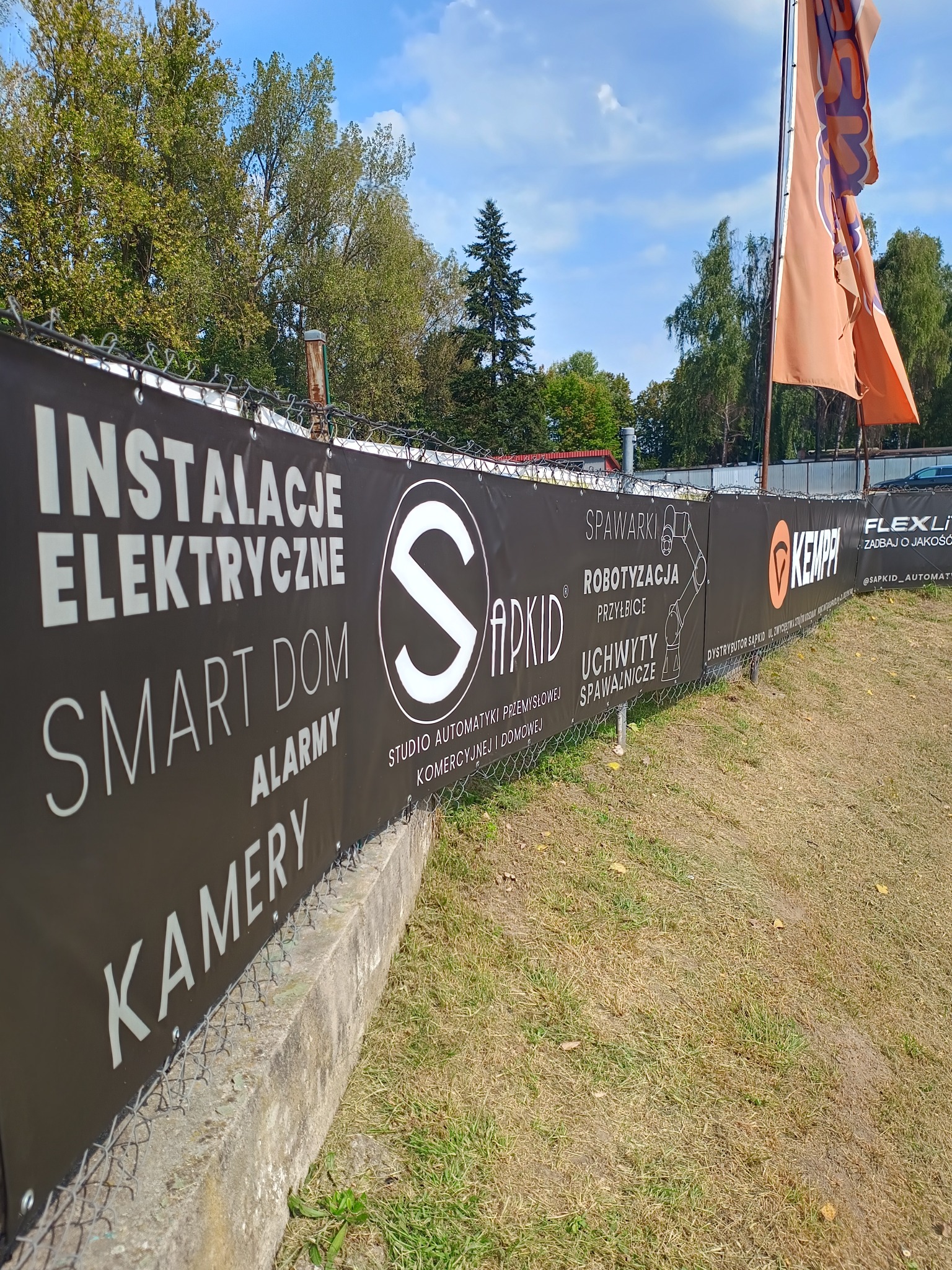Baner reklamowy firmy oferującej instalacje elektryczne, inteligentny dom, alarmy i kamery, umieszczony na ogrodzeniu z siatki, z widocznym logo firmy i informacjami o spawalnictwie.