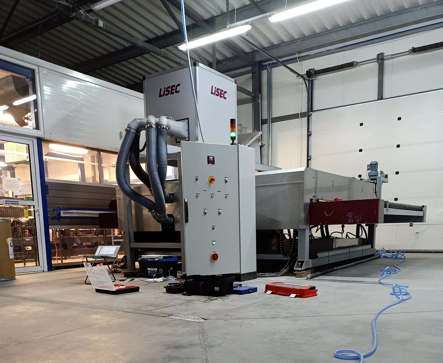 Duża maszyna przemysłowa marki LISEC w hali produkcyjnej, widoczna szafa sterownicza z przyciskami, przewody, skrzynki narzędziowe i otwarte pudełko z narzędziami na betonowej posadzce.