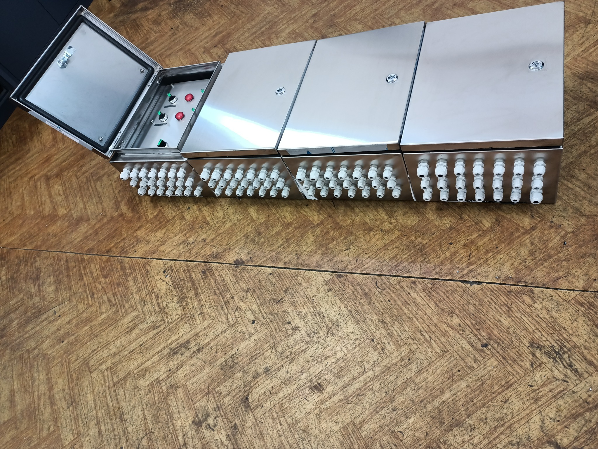 Cztery połączone metalowe skrzynki elektryczne z licznymi białymi dławikami kablowymi, jedna skrzynka otwarta ukazująca wewnętrzne przełączniki i diody, całość na drewnianej podłodze.