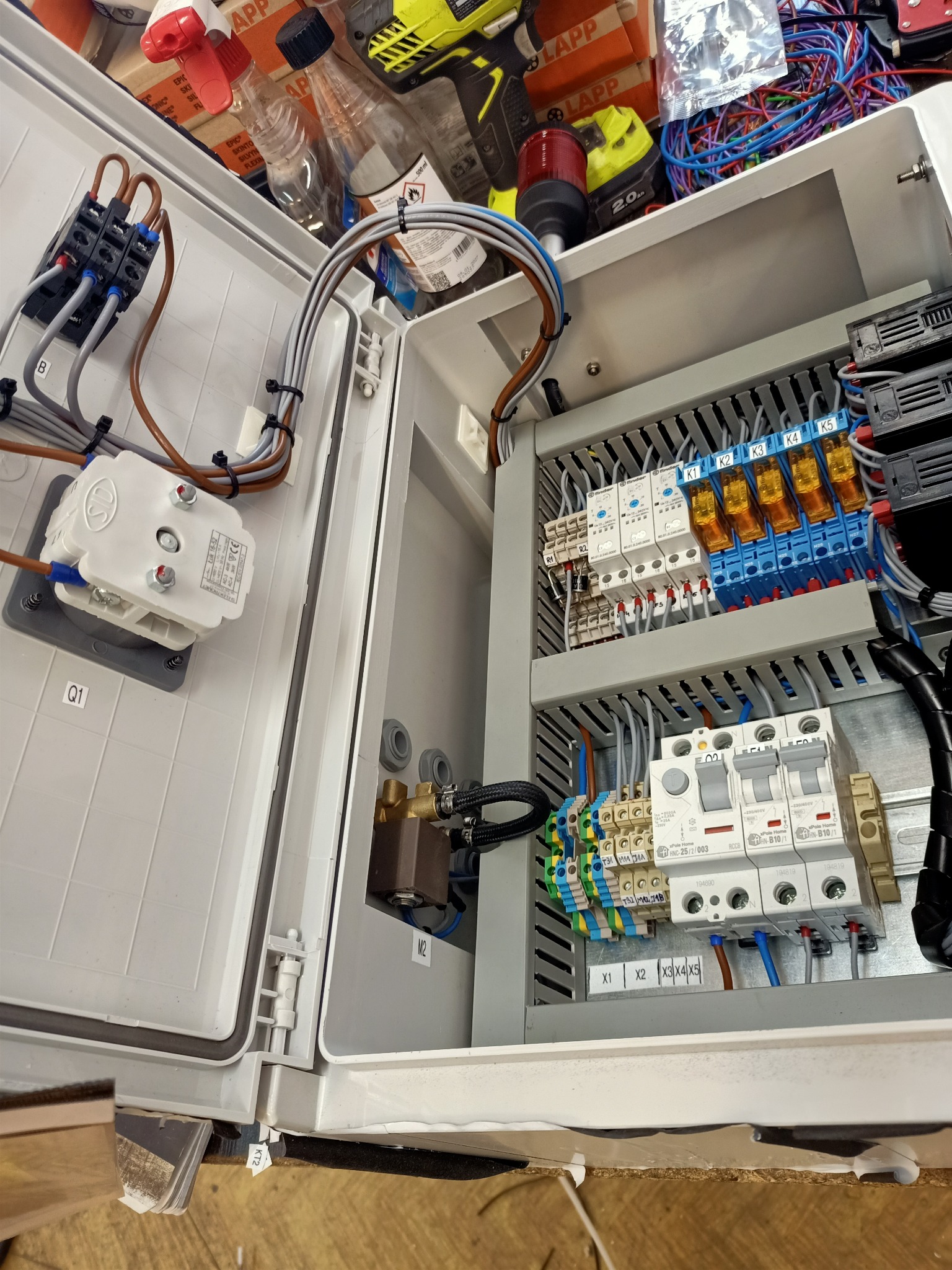 Otwarta szafa sterownicza z widocznymi elementami elektrycznymi: styczniki, bezpieczniki, przewody w peszlach, złączki, opisane etykietami, z narzędziami i akcesoriami na górze.