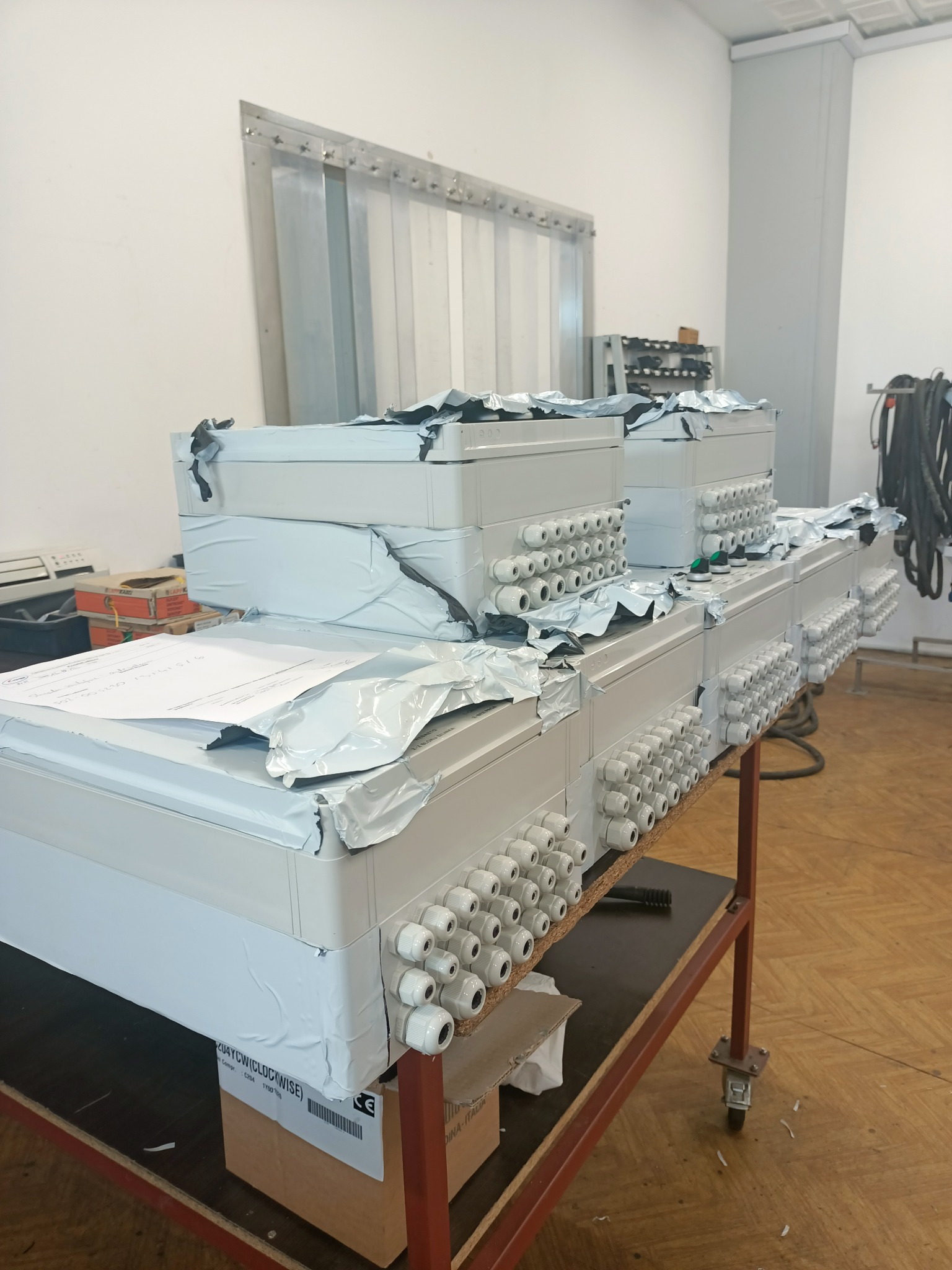 Przygotowane do montażu białe skrzynki elektryczne z licznymi dławikami kablowymi, częściowo owinięte folią, ustawione na wózku warsztatowym w pomieszczeniu.