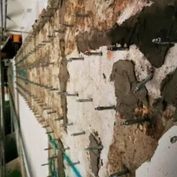 Zniszczona ściana z widocznymi śladami wilgoci i licznymi metalowymi kotwami w trakcie wzmacniania struktury. Fragmenty starego tynku i betonu uzupełnione świeżą zaprawą.