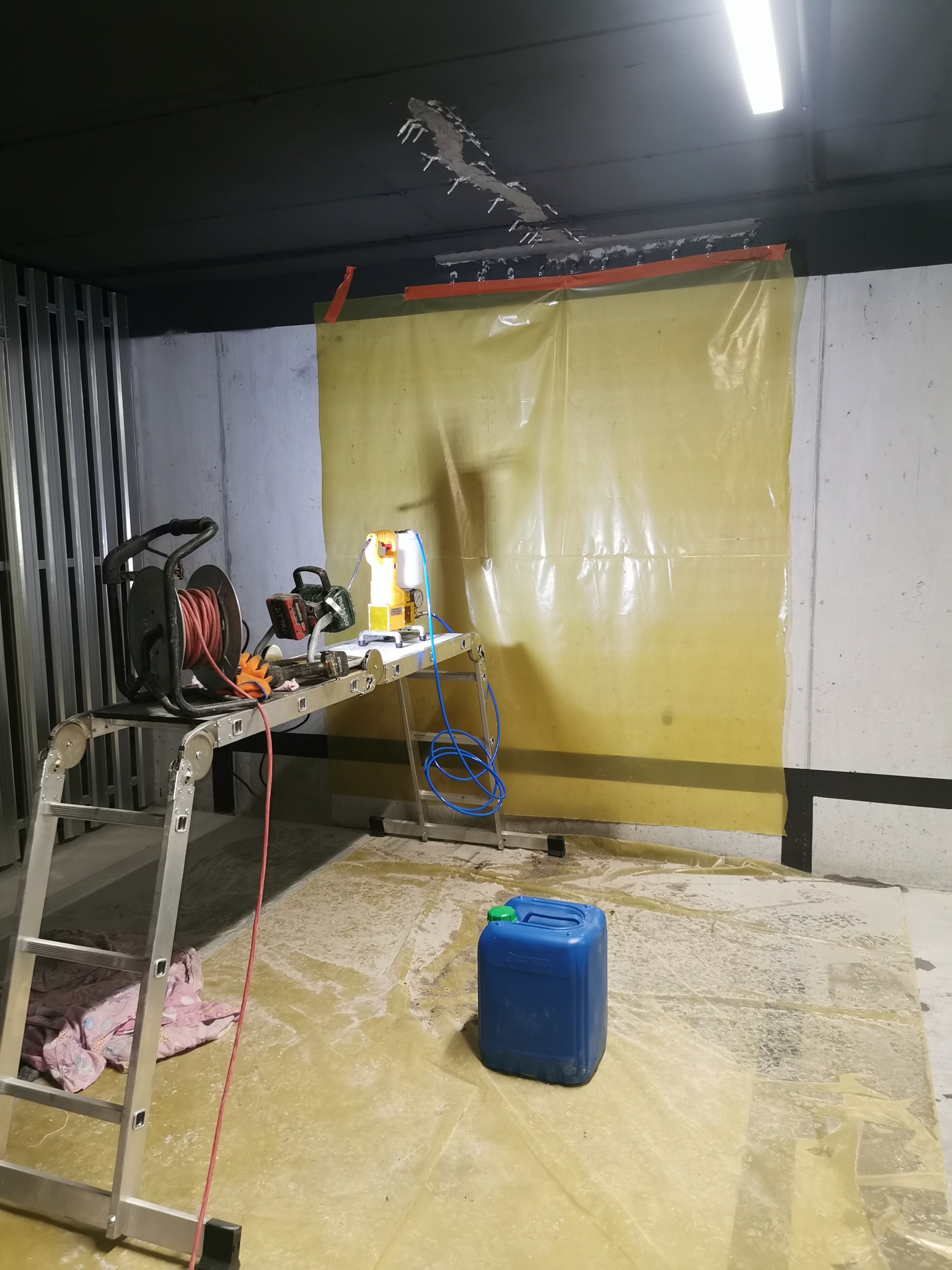 Przygotowania do prac: rozłożona drabina z przedłużaczem, agregat malarski na platformie, folia ochronna na ścianie i podłodze, niebieski kanister z płynem.