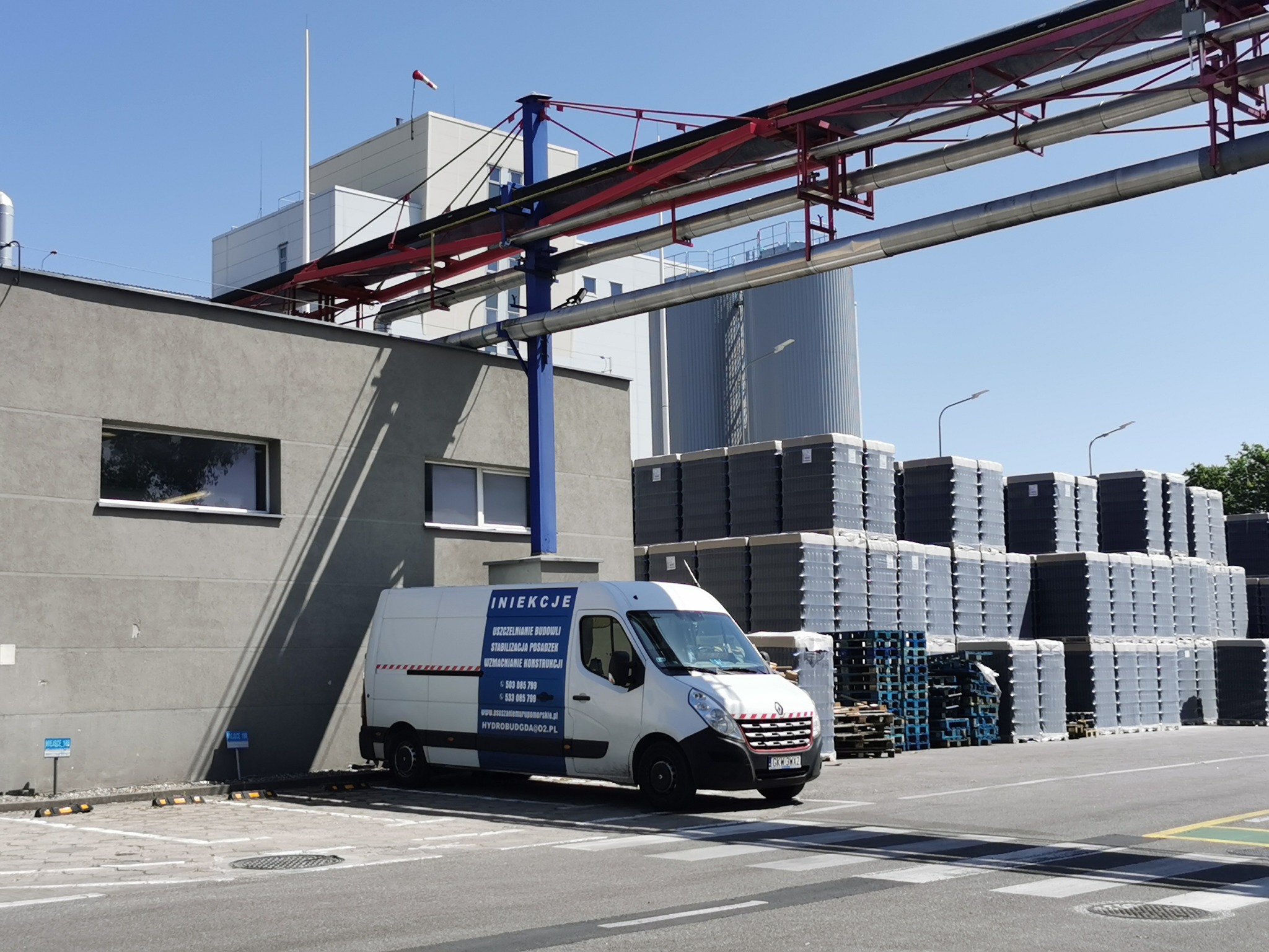 Biała furgonetka z reklamą usług iniekcji budowlanych zaparkowana na parkingu przemysłowym, w tle stosy szarych palet z materiałami budowlanymi i budynek przemysłowy z metalowymi rurami...