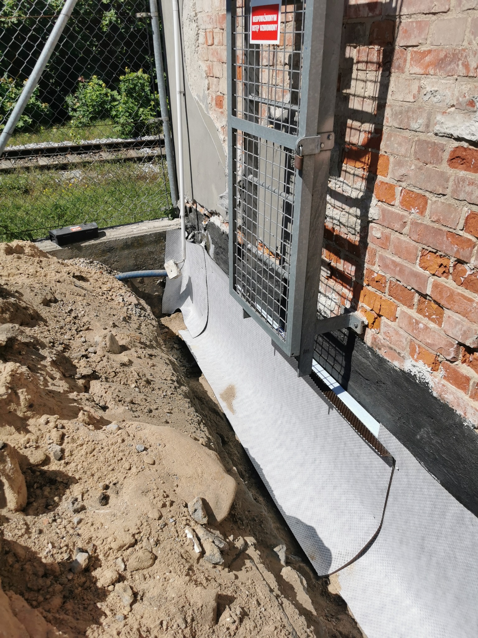 Odkopane fundamenty budynku z cegły, zabezpieczone białą membraną hydroizolacyjną i metalową kratą ochronną z tabliczką ostrzegawczą. W tle widoczna siatka ogrodzeniowa i tory kolejowe.