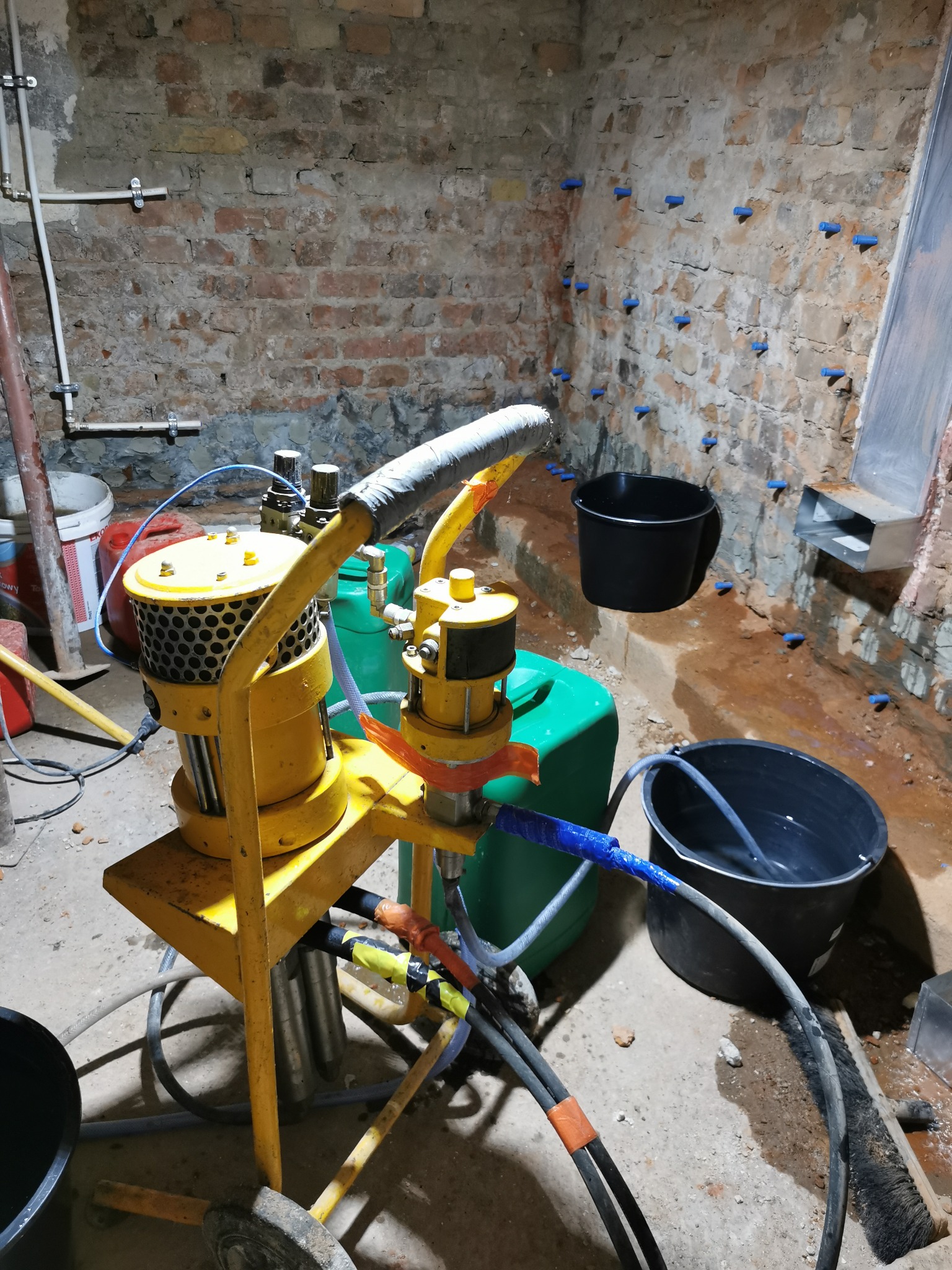 Żółta maszyna do iniekcji w wilgotnej piwnicy z ceglanymi ścianami i niebieskimi korkami iniekcyjnymi, obok stoją czarne wiadra i zielony pojemnik.