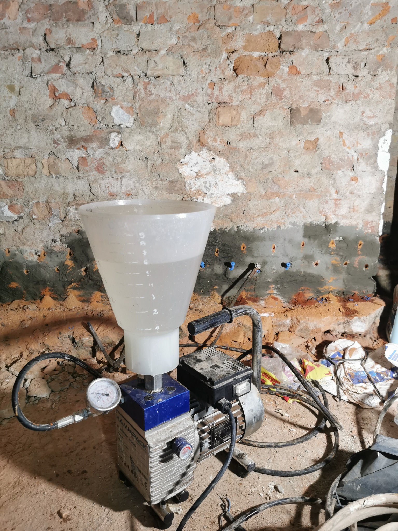 Urządzenie do iniekcji krystalicznej z przezroczystym zbiornikiem na płyn, widoczne nawiercone otwory w ceglanej ścianie z aplikatorami, manometr na obudowie pompy.