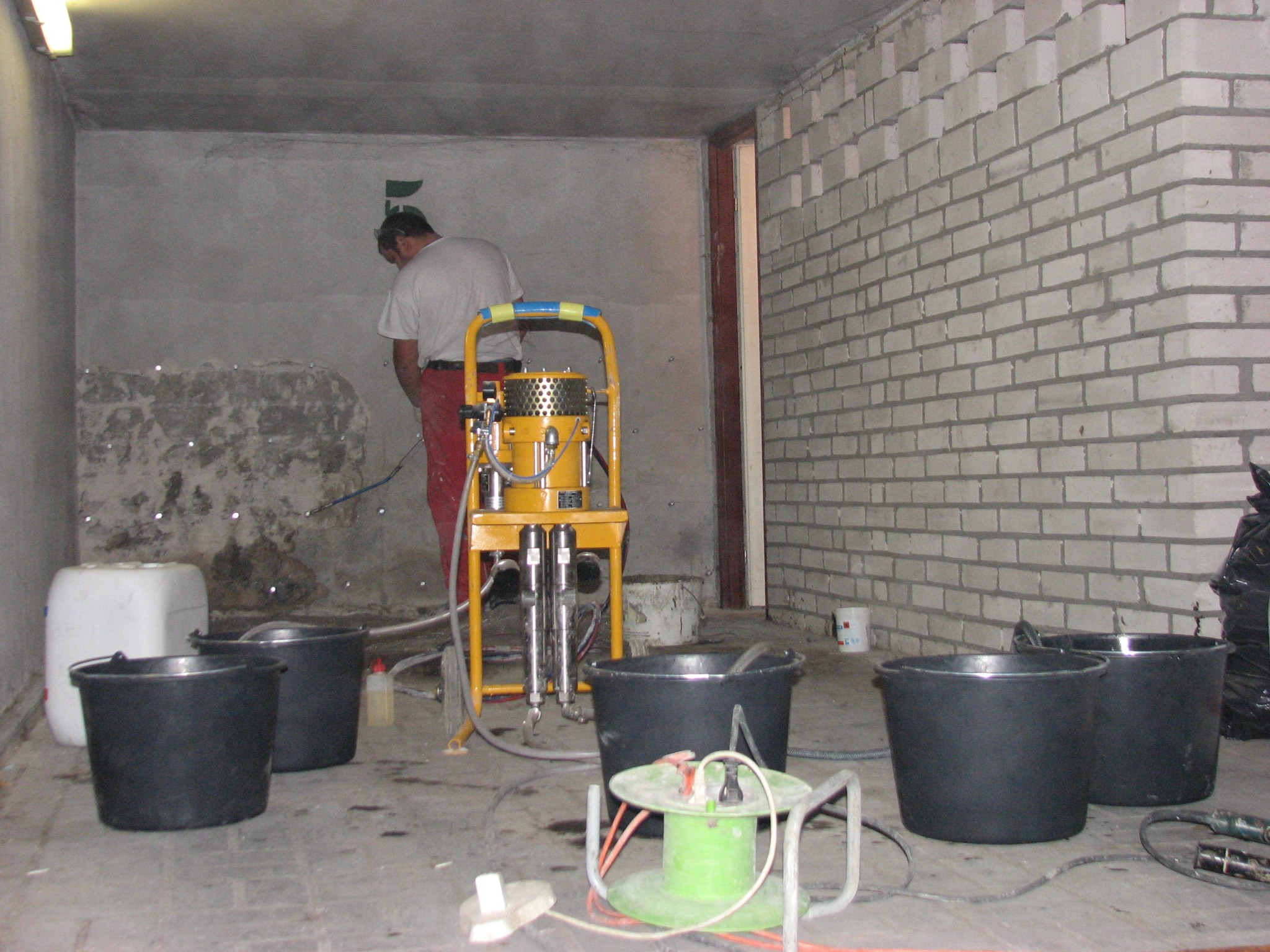 Pracownik w czerwonych spodniach obsługuje żółte urządzenie natryskowe w piwnicy z surowymi ścianami; widoczne wiadra, przedłużacz i ślady wilgoci na ścianie.