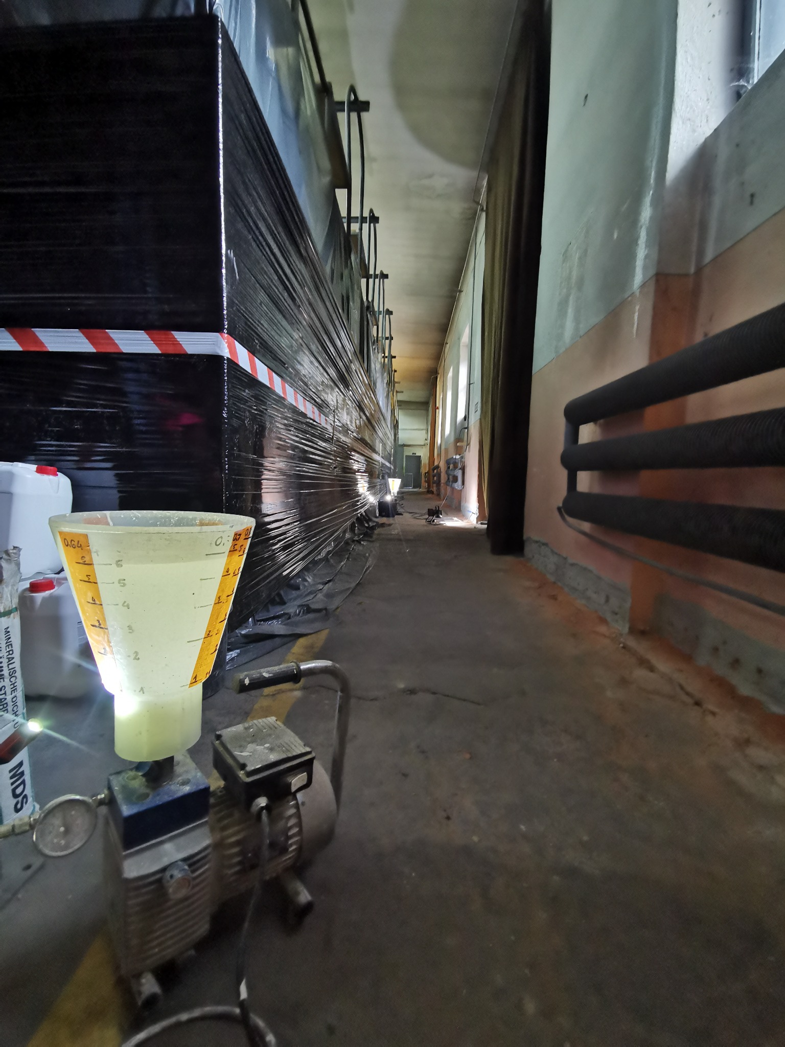 Sprzęt do pomiaru wilgotności z miarką i pojemnikami z płynem, na tle zabezpieczonej folią ściany z widocznymi śladami wilgoci w długim korytarzu.