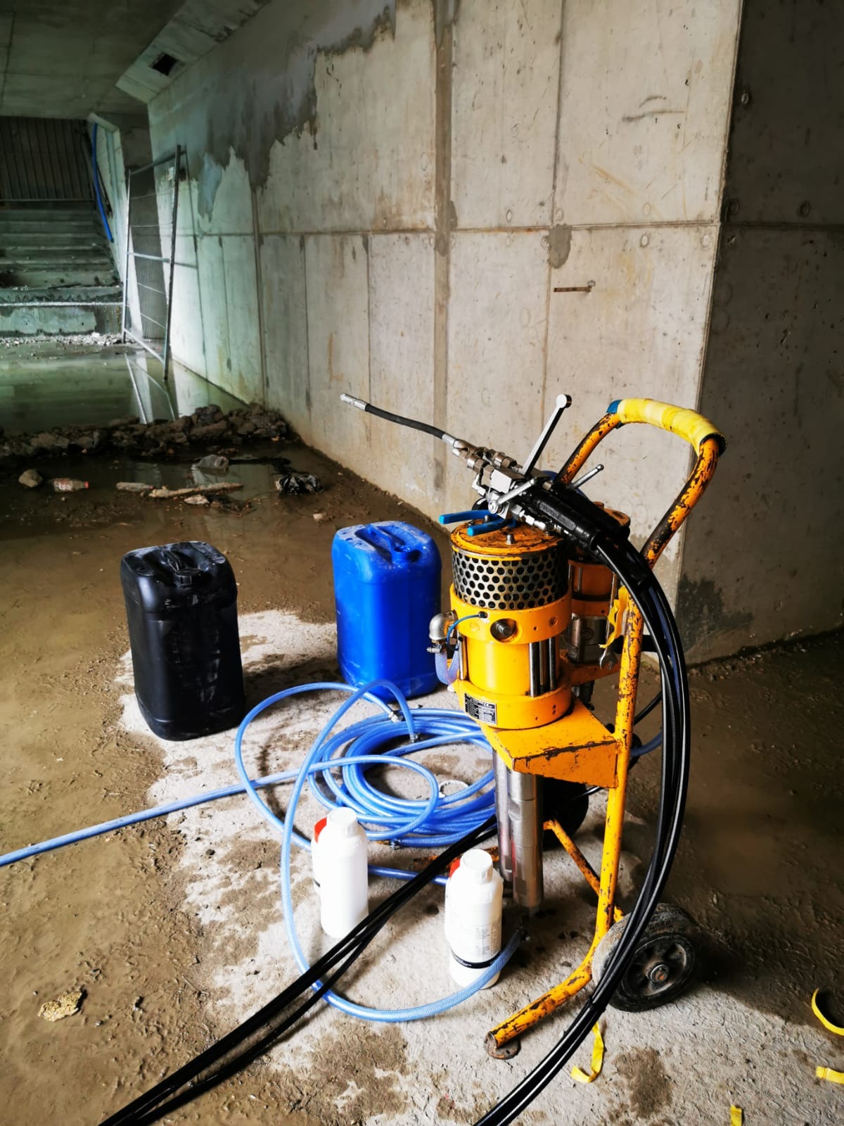 Żółty agregat iniekcyjny na wózku z niebieskimi wężami i kanistrami w betonowym pomieszczeniu z widoczną wodą na podłodze, prawdopodobnie w trakcie lub po pracy.