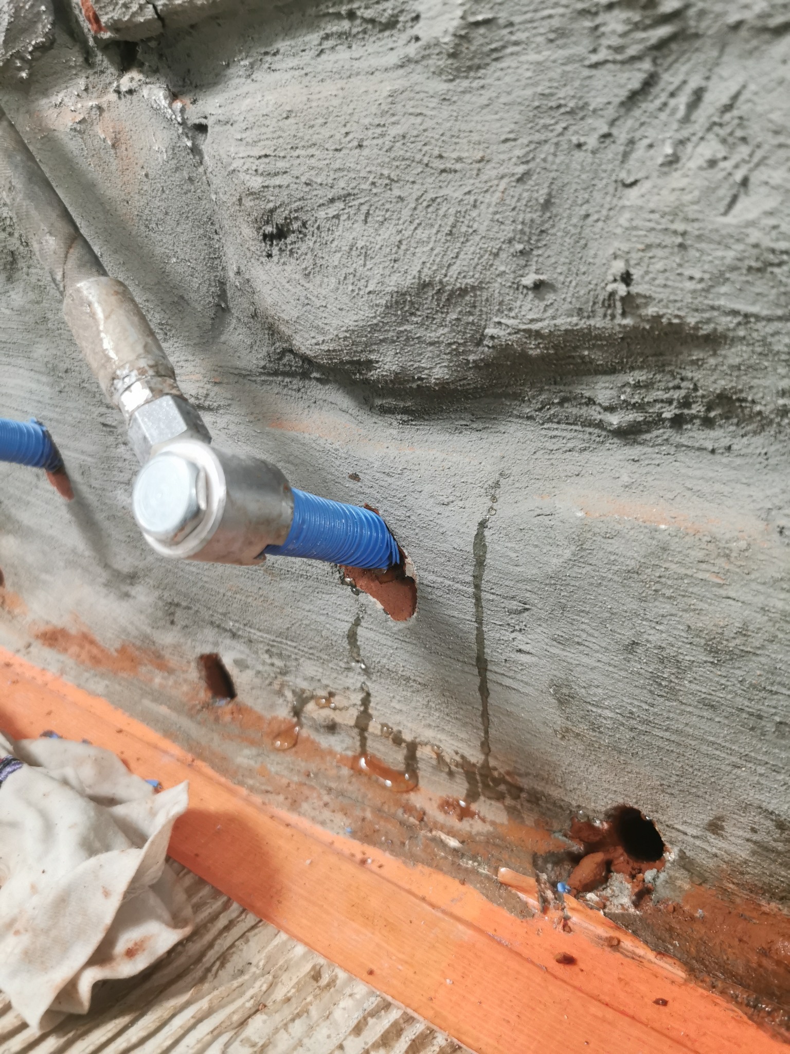 Mokry betonowy mur z niebieską rurą w metalowej obejmie, ślady wilgoci i plamy na ścianie, pomarańczowa listwa przypodłogowa i fragment zabrudzonej szmatki.