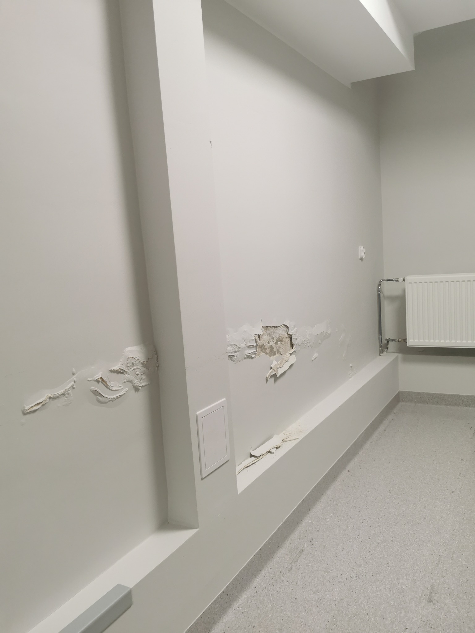 Biała ściana z widocznymi śladami wilgoci i odpadającą farbą, grzejnik na ścianie w tle, widoczny fragment podłogi w jasnych kolorach.