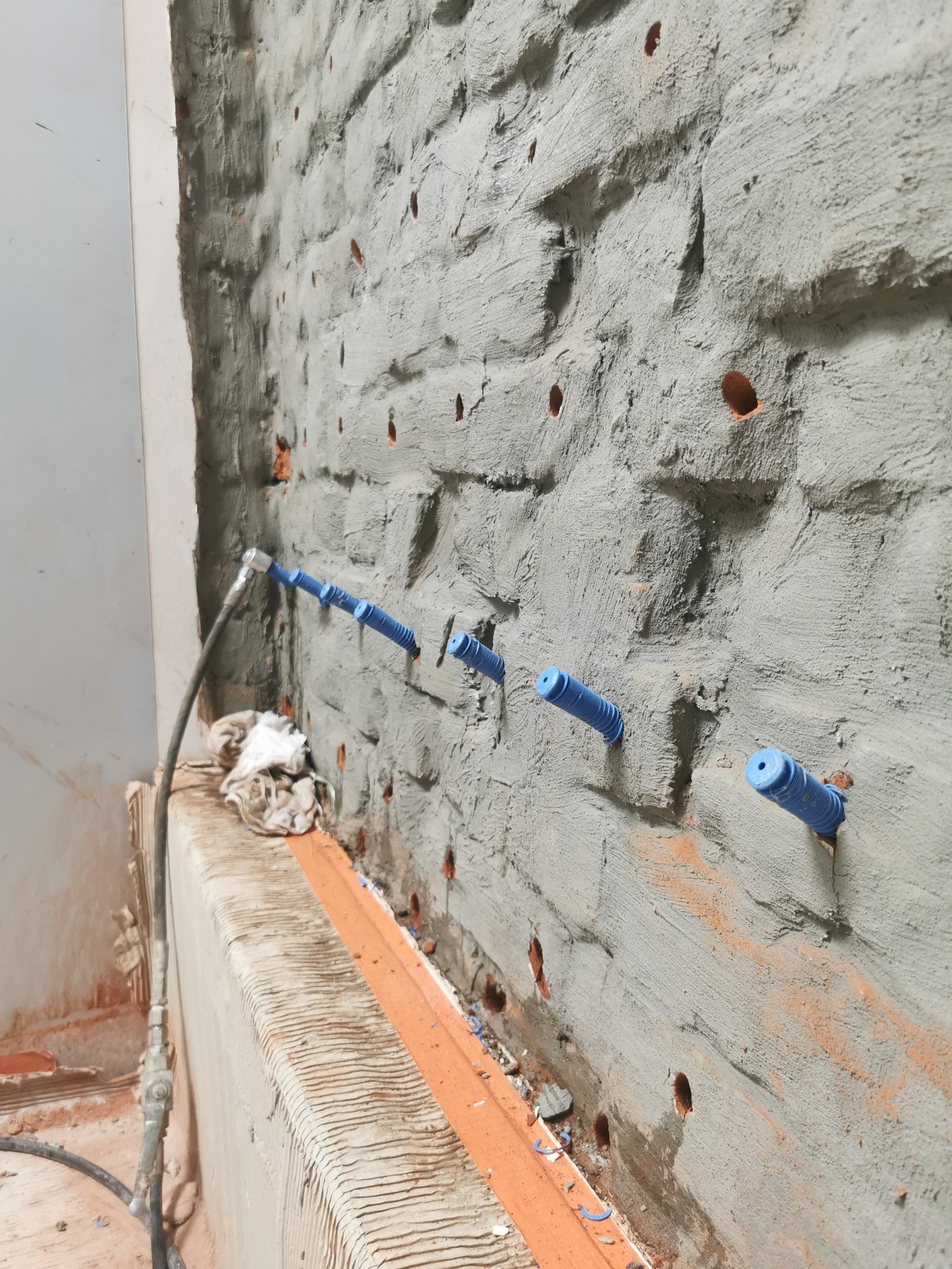 Świeżo otynkowana ściana z widocznymi otworami, w których umieszczono niebieskie, karbowane rurki do iniekcji; widoczny przewód iniekcyjny i zabrudzona szmatka.