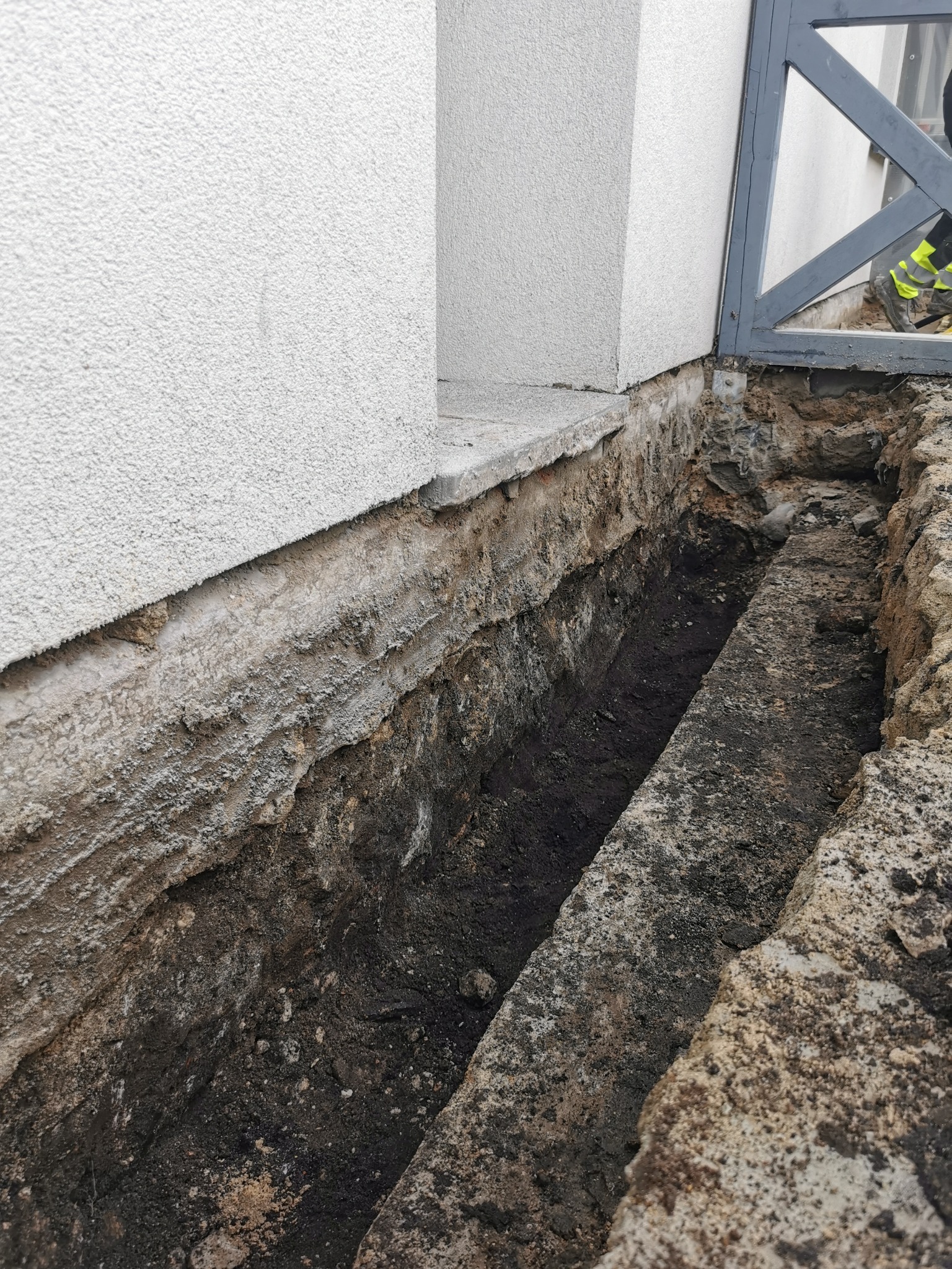 Wykop fundamentowy wzdłuż ściany budynku z widoczną izolacją przeciwwilgociową, częściowo odsłoniętą ziemią i fragmentem metalowej bramy w tle.