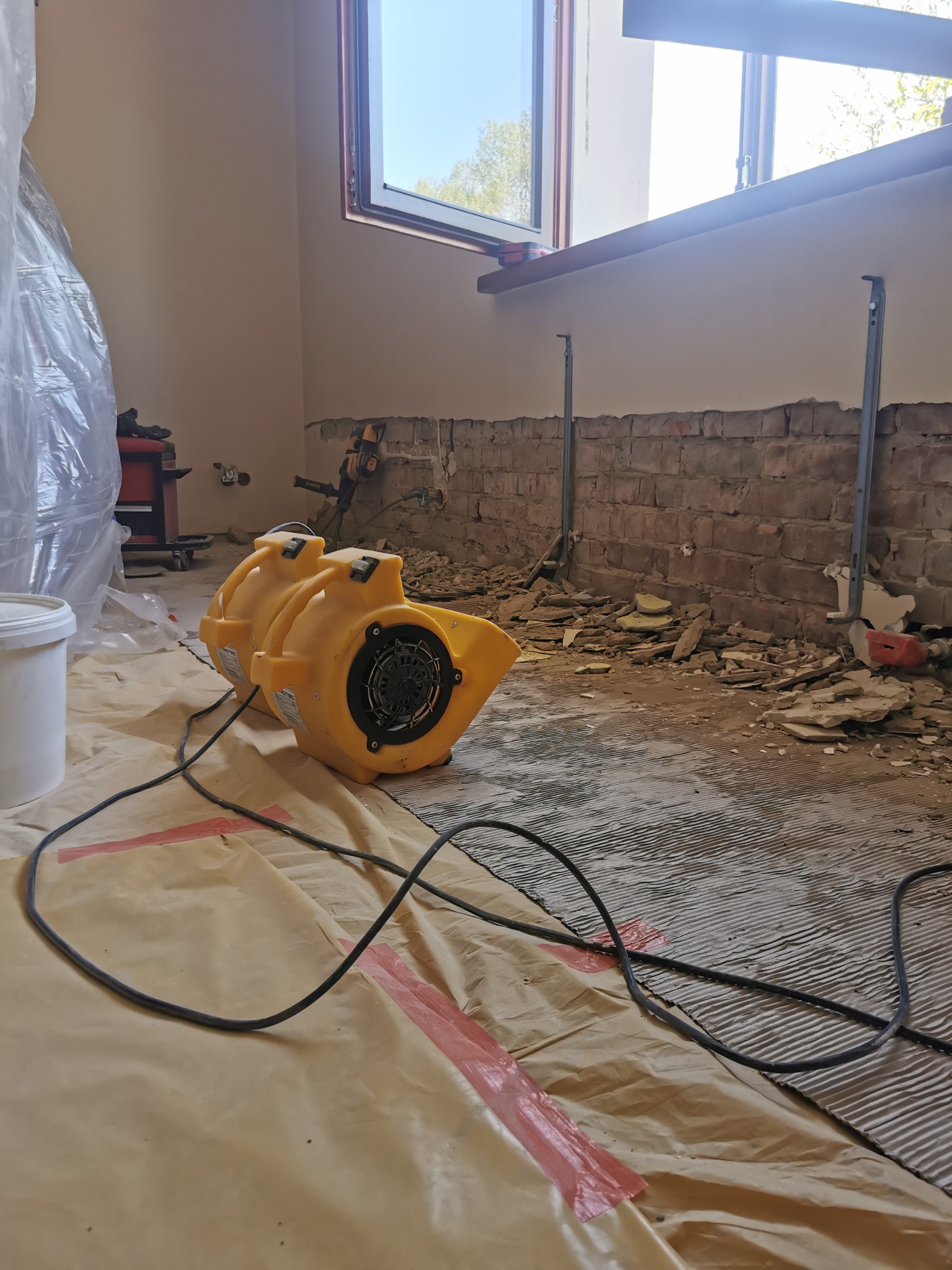Dwa żółte wentylatory przemysłowe na folii ochronnej w pomieszczeniu w trakcie remontu; widoczne fragmenty odsłoniętej cegły, gruz i narzędzia.