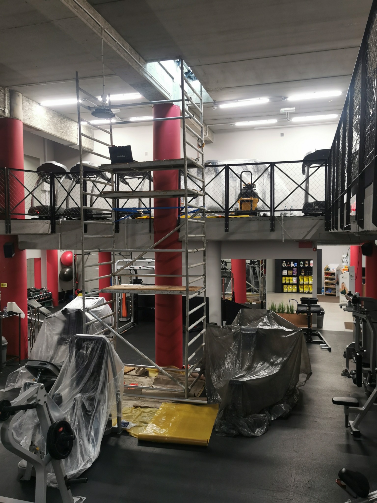 Wnętrze siłowni z rusztowaniem ustawionym wokół czerwonych kolumn, sprzęt częściowo zakryty folią ochronną, widoczny laptop na rusztowaniu.