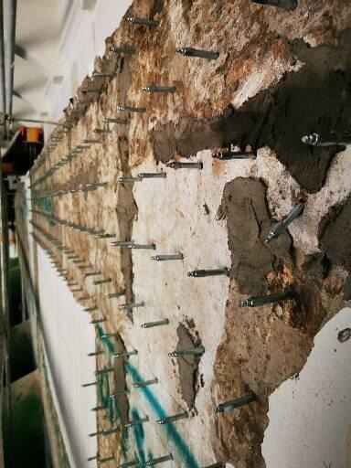 Zniszczona ściana z widocznymi śladami wilgoci i licznymi metalowymi kotwami w trakcie wzmacniania struktury. Fragmenty starego tynku i betonu uzupełnione świeżą zaprawą.