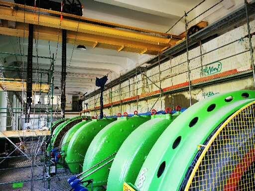 Wnętrze hali z rzędem dużych, zielonych turbin w trakcie prac remontowych, widoczne rusztowania i suwnica pod sufitem.