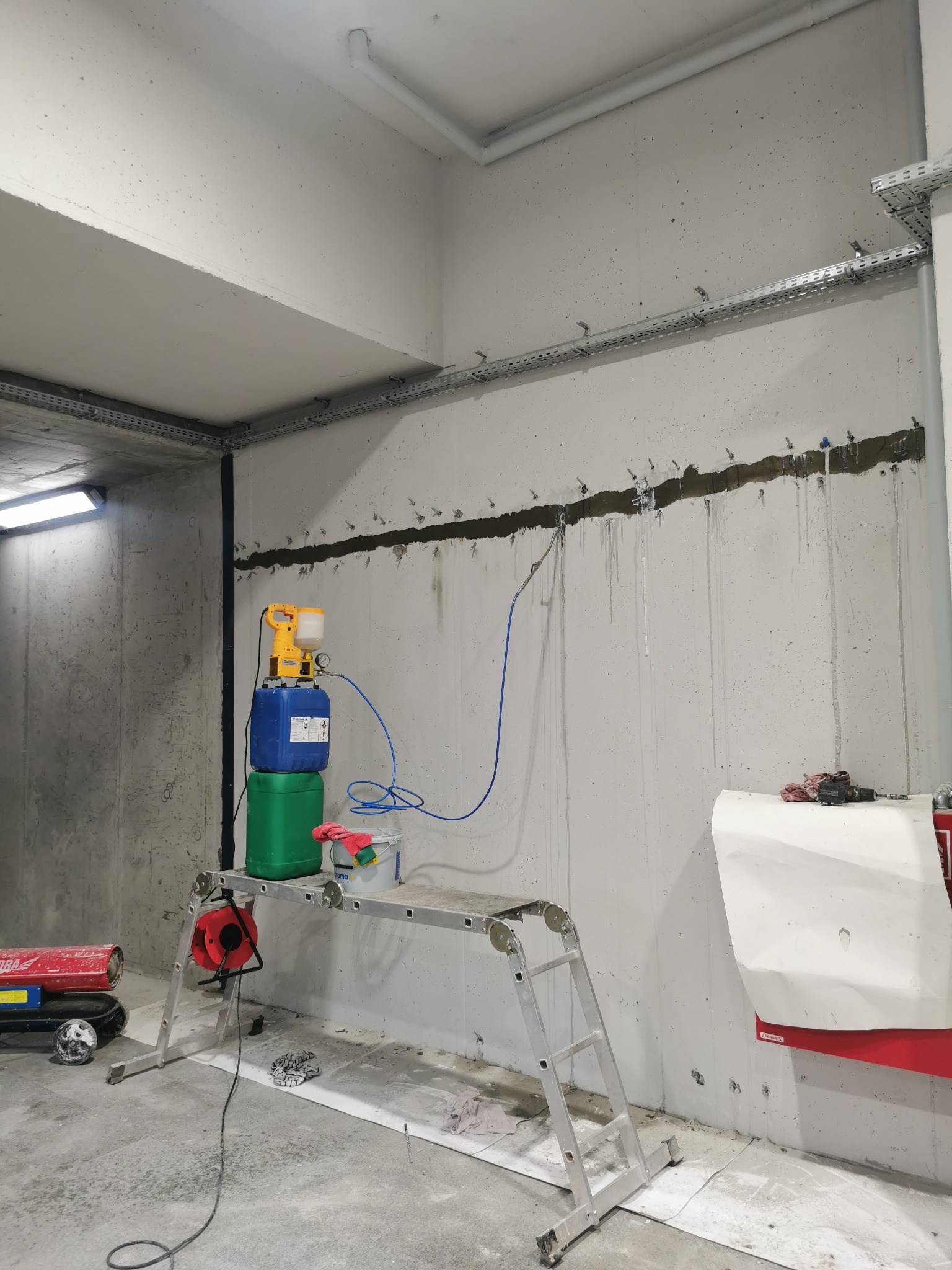 Sprzęt do iniekcji ciśnieniowej na podeście, widoczne ślady wilgoci na betonowej ścianie po wykonanej iniekcji, kable i rury pod sufitem.