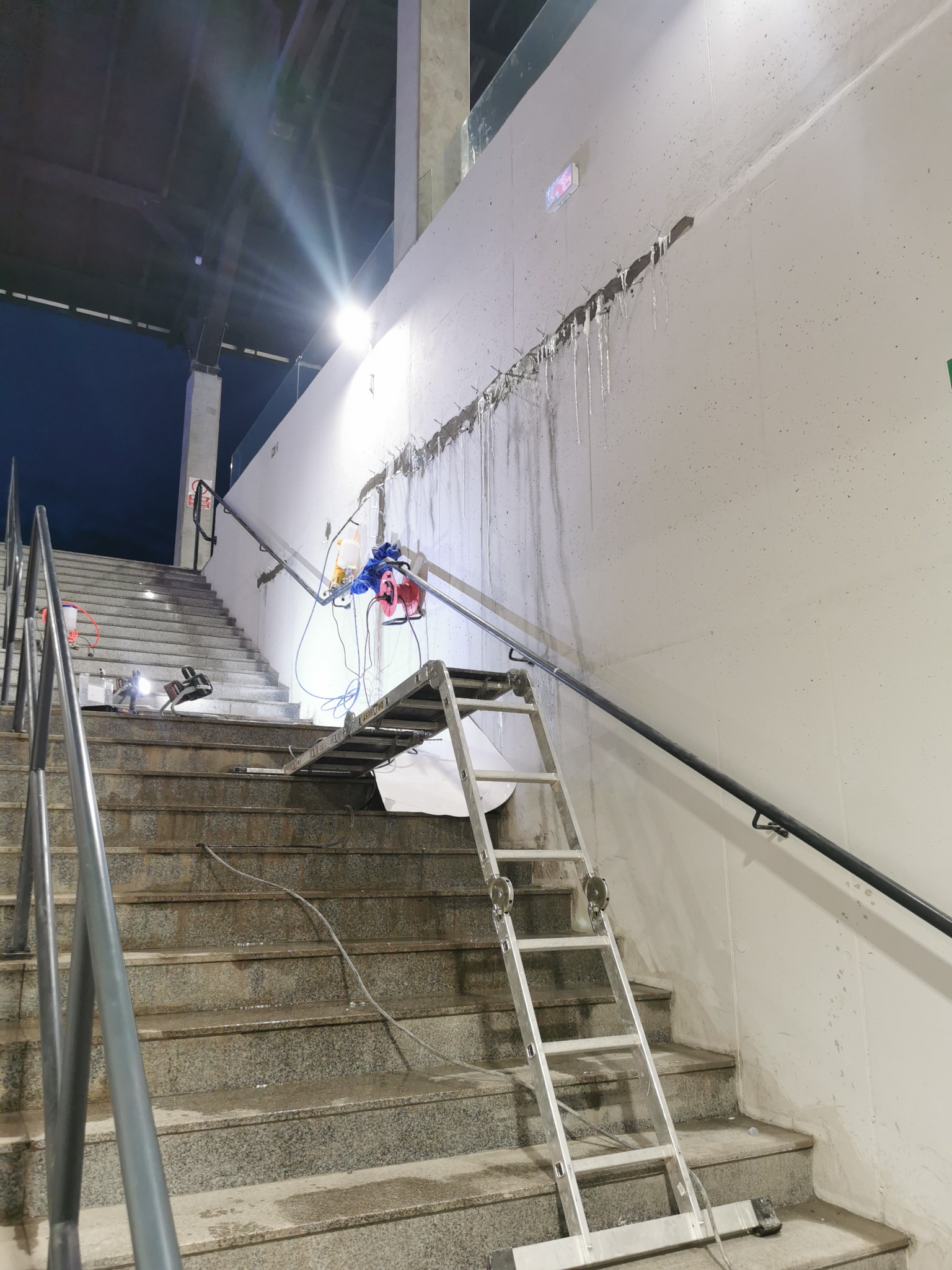Schody z marmurowymi stopniami, na których stoi aluminiowa drabina, a na ścianie widoczne ślady wilgoci i zamocowania sprzętu do osuszania, wskazujące na proces naprawczy.