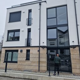 P&B Consulting Paweł Boniek - Trzypiętrowy budynek wielorodzinny z białymi ścianami i dużymi, czarnymi oknami, częściowo z roletami zewnętrznymi, widoczny przez metalowe ogrodzenie od strony chodnika, z mężczyzną stojącym przed...